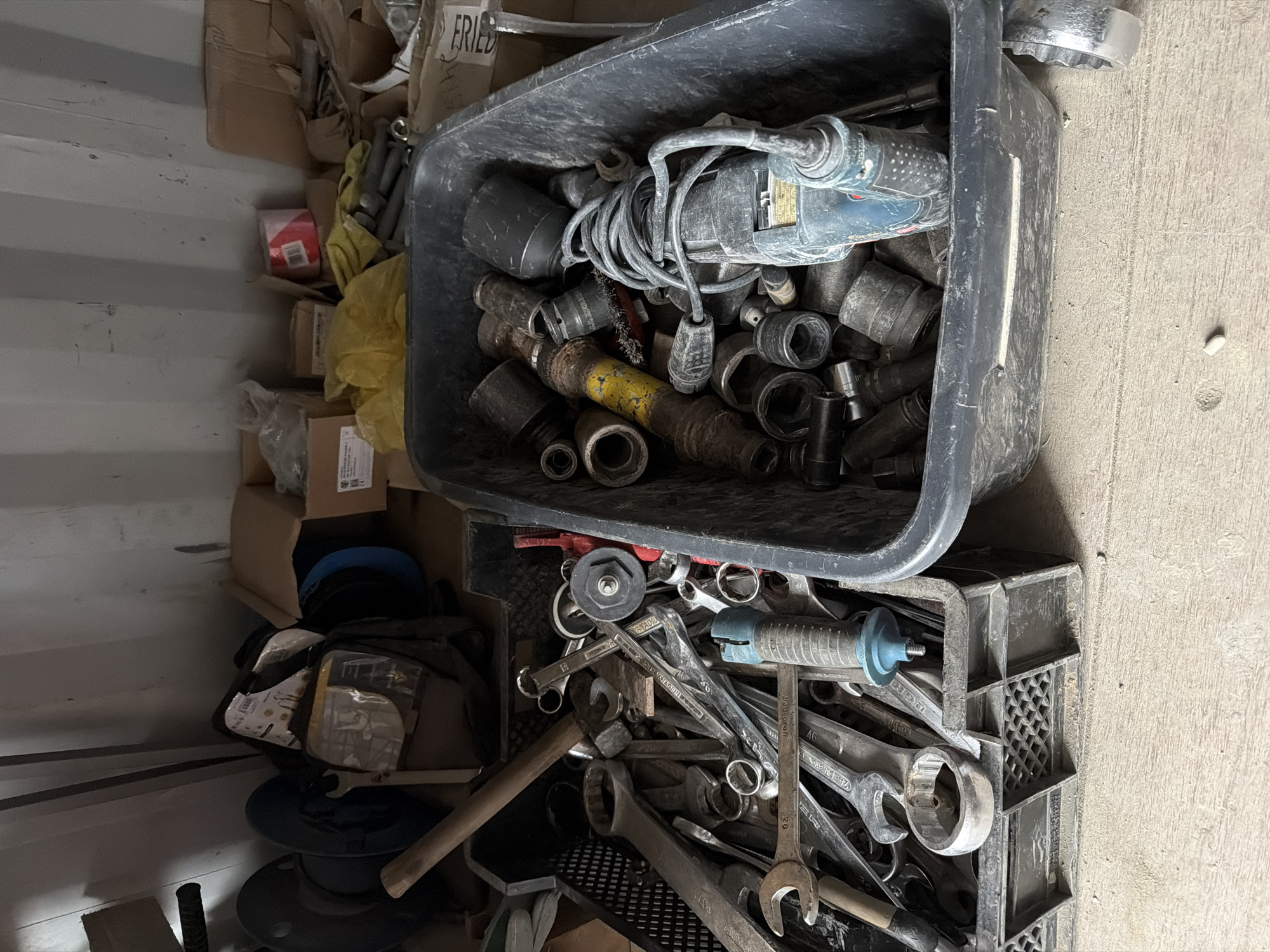 This image depicts a cluttered storage area, likely a workshop or garage, within the city of Janowice, Poland. The primary focus is on two large, dark-colored bins overflowing with tools and hardware. The bin on the left contains a variety of wrenches, including large open-end and box-end wrenches, as well as some smaller tools and a wooden-handled tool. This bin is a black plastic crate with a grid pattern. The bin on the right, which appears to be a wheelbarrow or a large plastic tub, is filled with various socket wrenches, extensions, and an electric impact wrench. The impact wrench, a blue and black Bosch model, is coiled with its power cord. Scattered amongst the tools are some cardboard boxes, a yellow plastic bag, and what appears to be a roll of red and white tape. The environment is dimly lit, with shadows cast from above, suggesting an indoor space with limited natural light. The floor appears to be concrete or a similar industrial material, with dust and debris scattered about. No people are visible in the image. The overall impression is one of disorganization and a functional, working space.