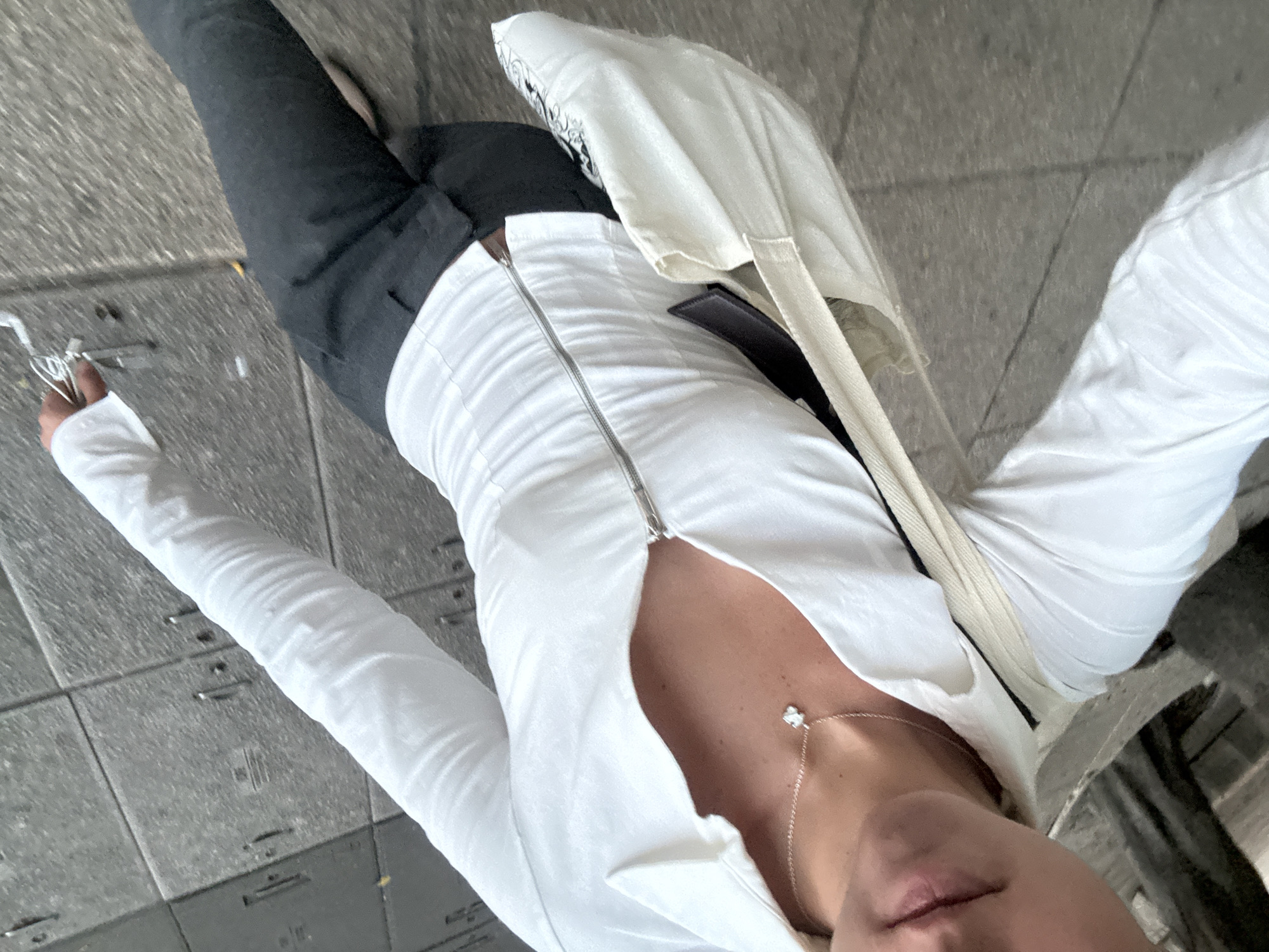 A top-down, full-body shot captures a person standing on a gray concrete sidewalk, likely in an urban setting. The person is wearing a white, long-sleeved top with a front zipper, dark pants, and a cream-colored tote bag slung over their shoulder. A delicate necklace is visible around the person's neck. In their left hand, they hold a small, unclear object. The perspective is slightly angled, giving a distorted view of the person's lower body and legs. The background shows the typical texture of a city sidewalk. The location is likely in Mexico City, Mexico.