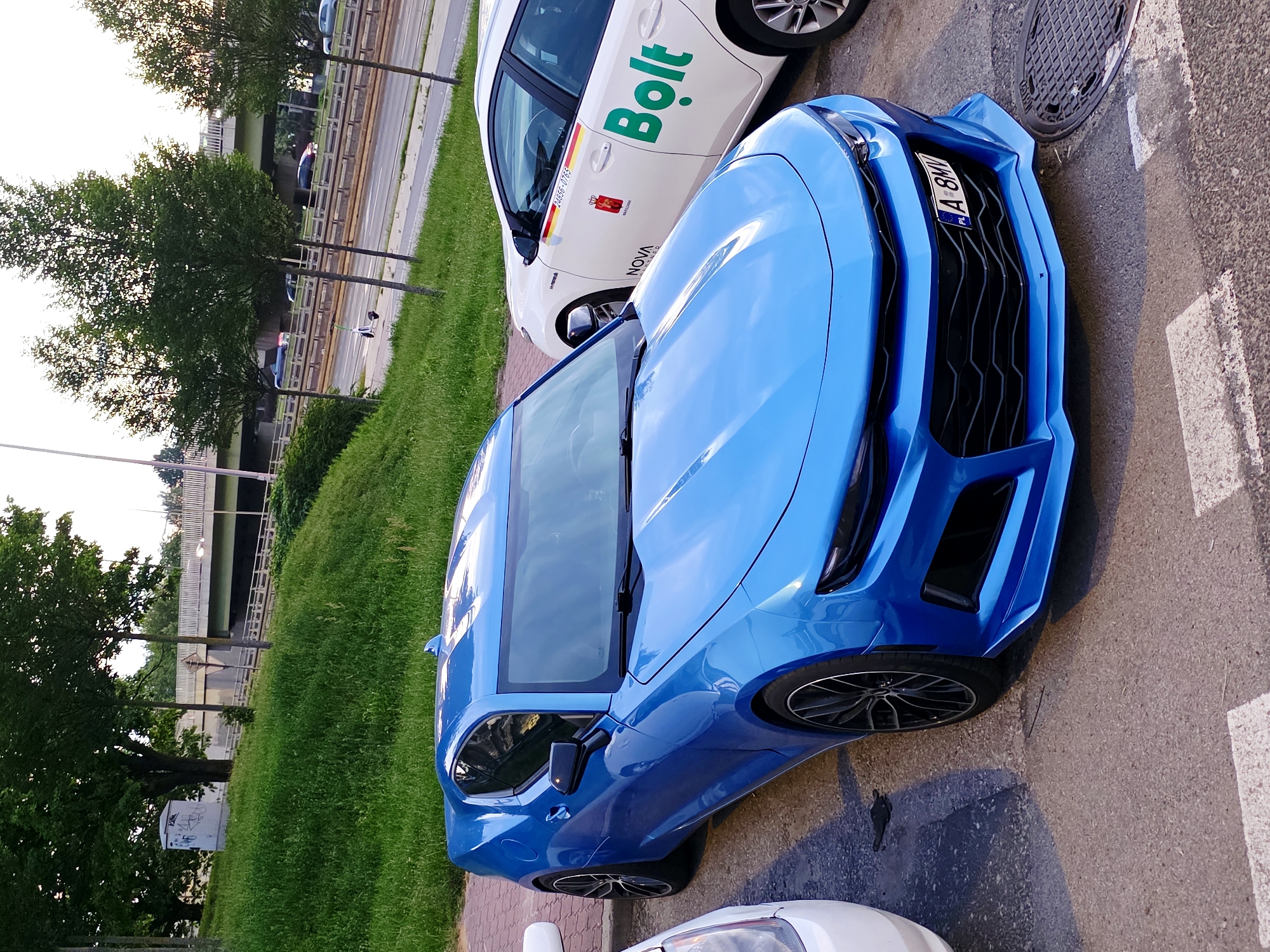 A blue Chevrolet Camaro is parked on a street in Warsaw, Poland, next to a white Bolt taxi. The Camaro is in the foreground, facing towards the right side of the frame. It has a sporty design with sharp lines and a large grille. The Bolt taxi is parked to its right, displaying the Bolt logo in green. In the background, there is a grassy embankment with trees and a highway overpass. Traffic can be seen on the highway. The sky is overcast.