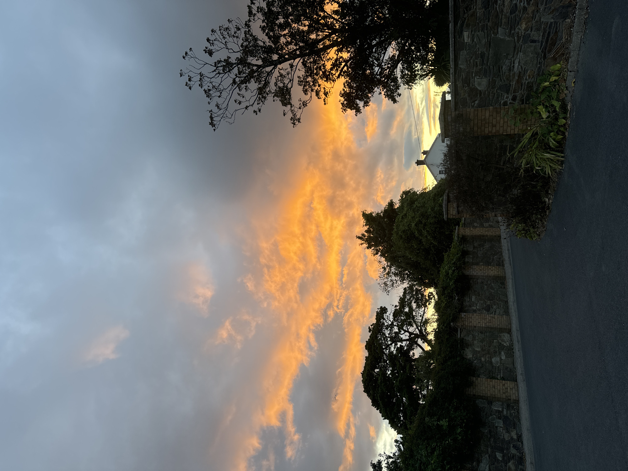 The image shows a road leading past a stone and brick wall under a dramatic sunset sky. The sky is filled with clouds tinged with vibrant orange and yellow hues, indicating either sunrise or sunset. Silhouetted trees stand against the colorful sky, and a portion of a white house with a chimney is visible behind the wall. The wall itself is constructed from irregular stones and interspersed with brick pillars. The road in the foreground is paved and appears empty of people or vehicles. The scene is likely located in a suburban or rural area, possibly on the outskirts of Waterford City, Ireland, given the stone wall construction and the presence of mature trees.