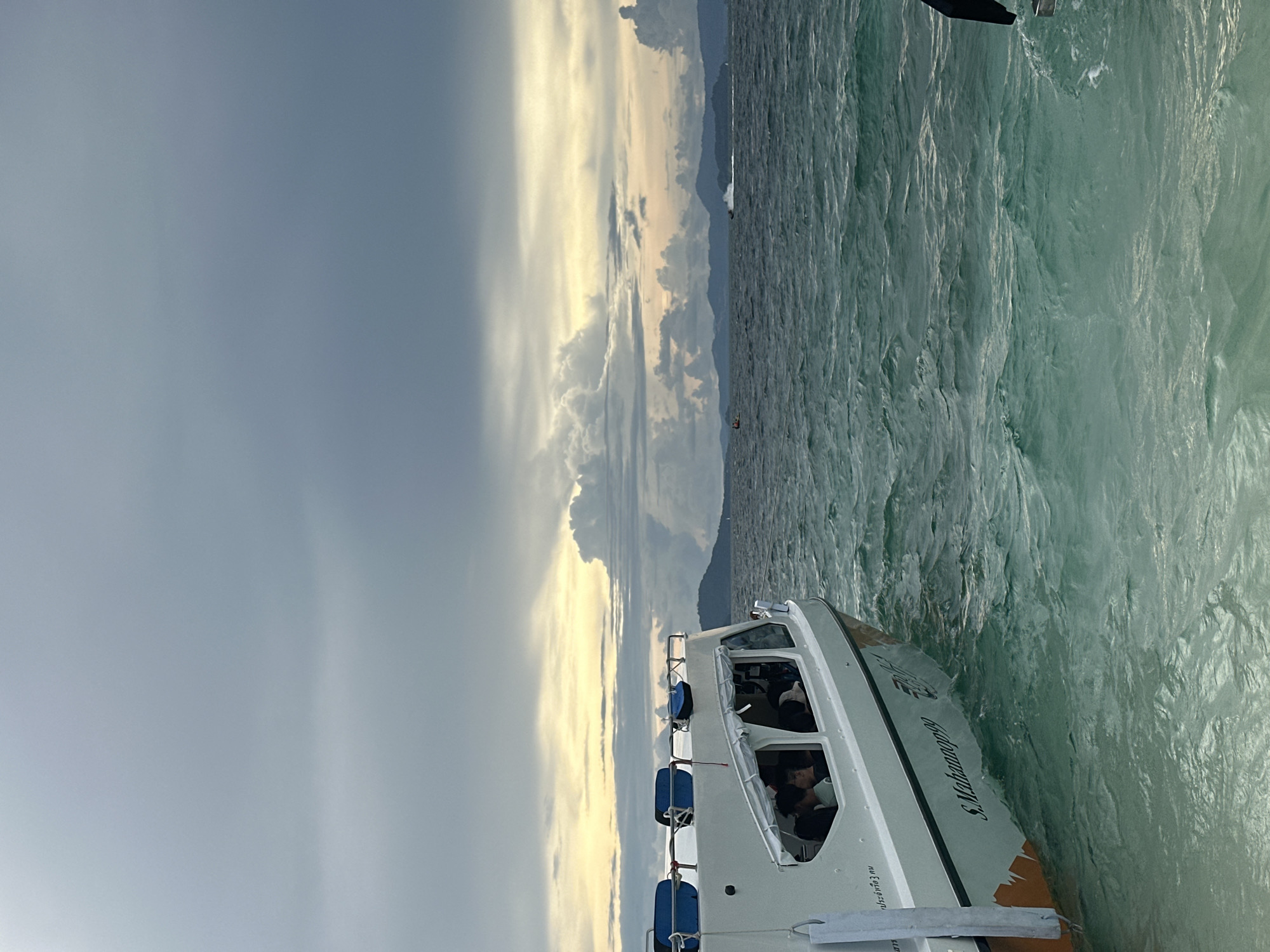 A white passenger boat with blue pontoons on top, labeled "S. Mahanop999" in gray text, is partially visible on the left side of the frame. The boat is in motion on choppy, turquoise water. Several passengers are visible inside the boat through the windows. In the distance, a hazy mountain range is visible under a dramatic, cloudy sky with patches of yellow light breaking through. Other small boats and what appears to be a jet ski are also present on the water in the distance. The scene appears to be along the coast of Phru Nai, Thailand.