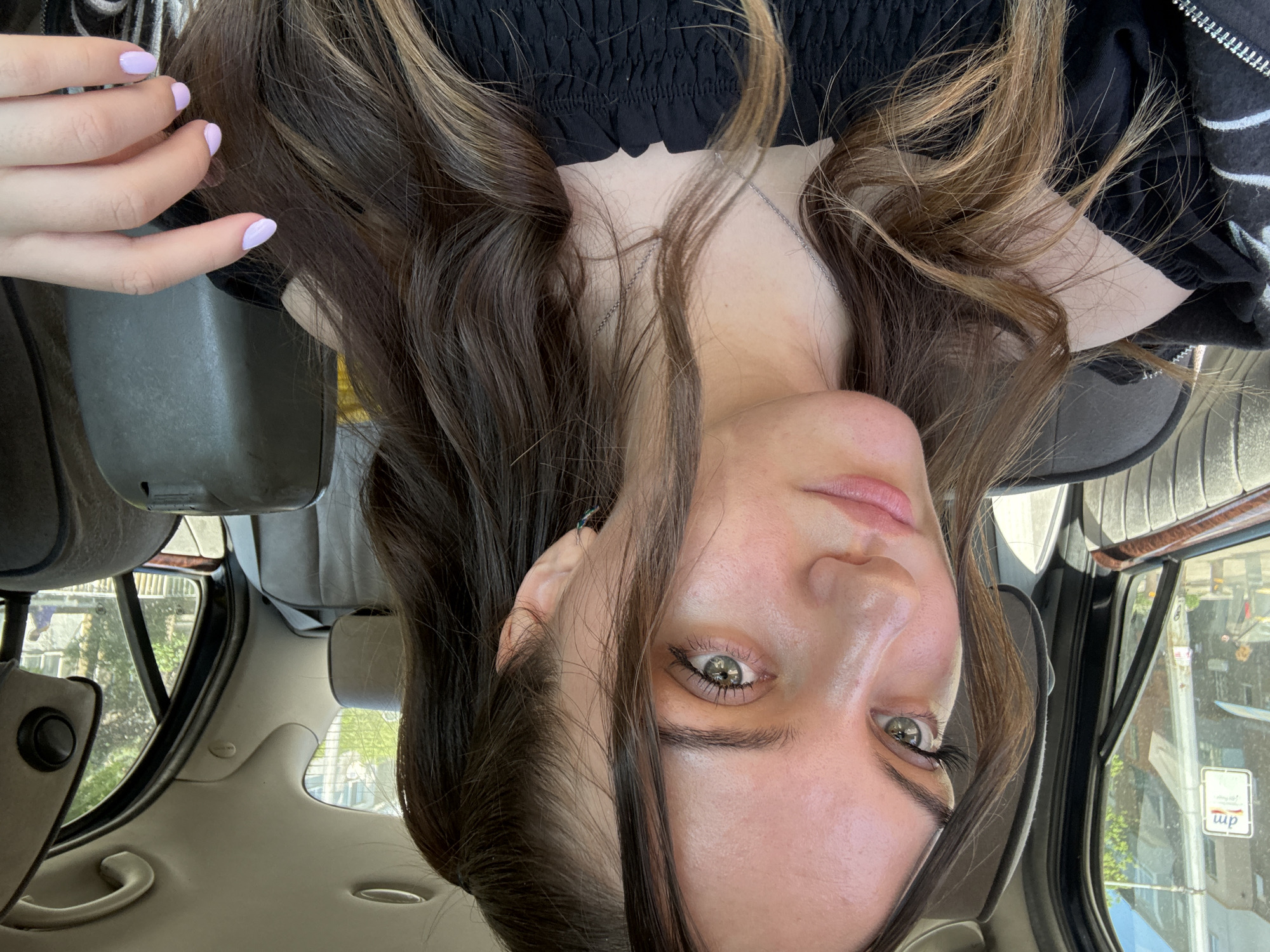 A young woman is inside a car, likely a vehicle. Her face is directed towards the camera, but the image is rotated upside down. She has long brown hair with blonde highlights, light skin, and green eyes. She is wearing a black top. Her left hand is visible in the upper left corner of the image with light purple nail polish. The interior of the car is visible. A portion of the car window shows a street scene outside, which is blurry but includes buildings and trees. There are also some decals on the window including one that reads "wp". Due to the decal on the side of the window, this photograph was likely taken in Sofia, Bulgaria.