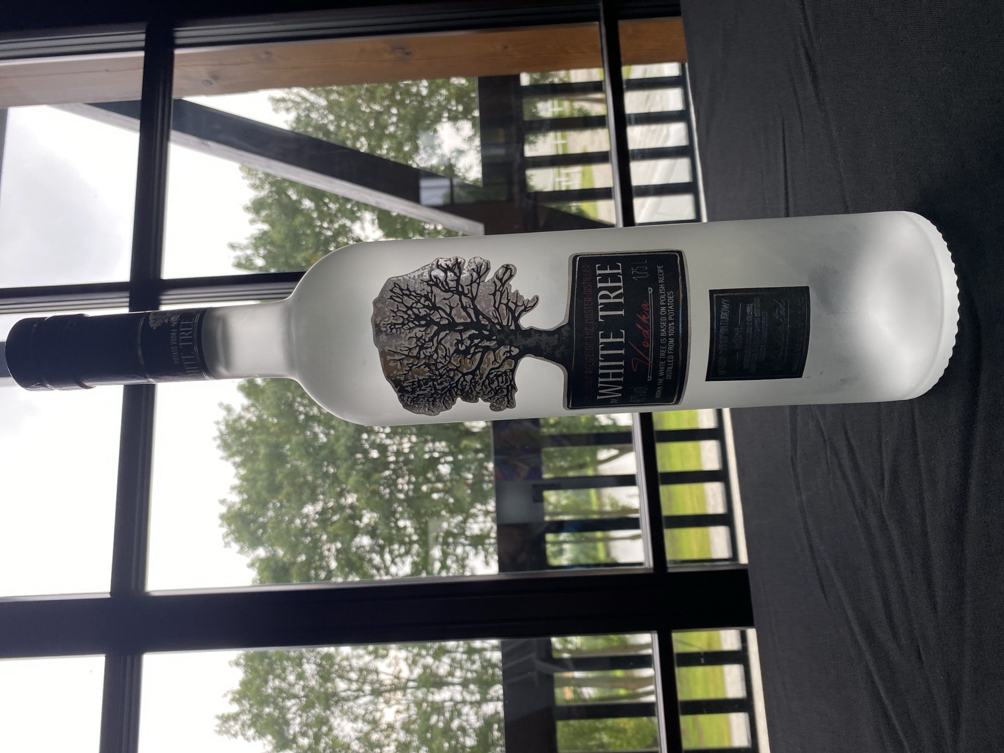 A frosted glass bottle of White Tree Vodka stands upright on a dark, textured surface, likely a tablecloth. The bottle has a black cap and a label featuring a silhouette of a tree and the text "WHITE TREE VODKA". Behind the bottle, a large window offers a view of lush green trees and a cloudy sky. The window frame is dark, with wooden beams and railings visible outside, suggesting an outdoor or semi-outdoor setting. The overall scene is calm and well-lit, with the bottle being the clear focal point. The presence of a Polish recipe mentioned on the label strongly implies the location is within Poland, and the characteristic architecture of the window and outdoor structures suggests a context in or near the city of Buk.