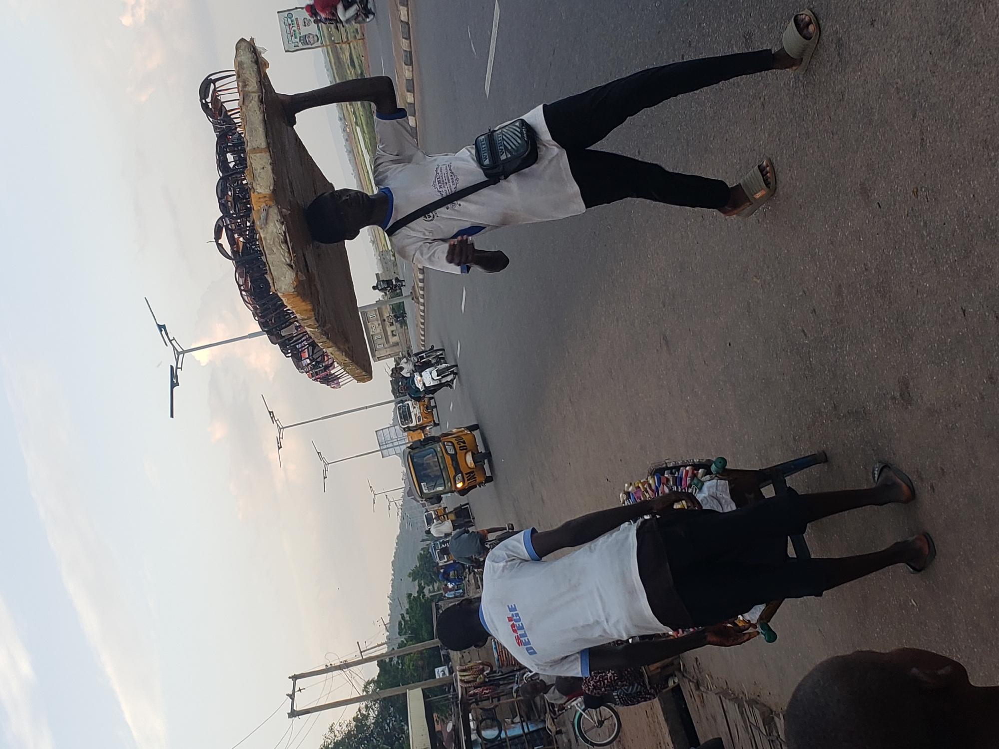 The image captures a vibrant street scene in Lokoja, Nigeria, depicting daily life and commerce under an overcast sky.
In the foreground, a man is prominently featured, skillfully balancing a large, flat, rectangular tray on his head. The tray is laden with numerous small, dark items, possibly hair accessories or other small merchandise, suggesting he is a street hawker. He wears a white t-shirt with blue trim, black trousers, and light-colored slip-on sandals. A small black shoulder bag, emblazoned with the word "WHITE," hangs across his chest. His t-shirt also features a crest and text that includes "KNOWLEDGE IS POWER," although not fully legible.
Further along the road, another vendor is visible, standing behind a small, blue-framed pushcart. This cart displays an array of colorful small items, likely cosmetics or toiletries, neatly arranged for sale. This vendor also wears a white t-shirt with blue accents, with text that appears to read "SALE DELEGE" or "SALES DELEGATE," along with dark shorts or trousers and dark sandals.
The background reveals a busy urban thoroughfare populated by various vehicles, including several distinctive yellow tricycles (commonly known as Keke Napep in Nigeria), motorcycles, and cars. Streetlights, utility poles with wires, and buildings line the road. In the far distance, hills are visible, and what appears to be a body of water, consistent with the River Niger or Benue given Lokoja's location, stretches across the horizon. A large billboard with faces and indistinct text, possibly advertising, can also be seen in the distance. The overall scene reflects the dynamic environment of a Nigerian city, with vendors plying their trade amidst everyday traffic.
