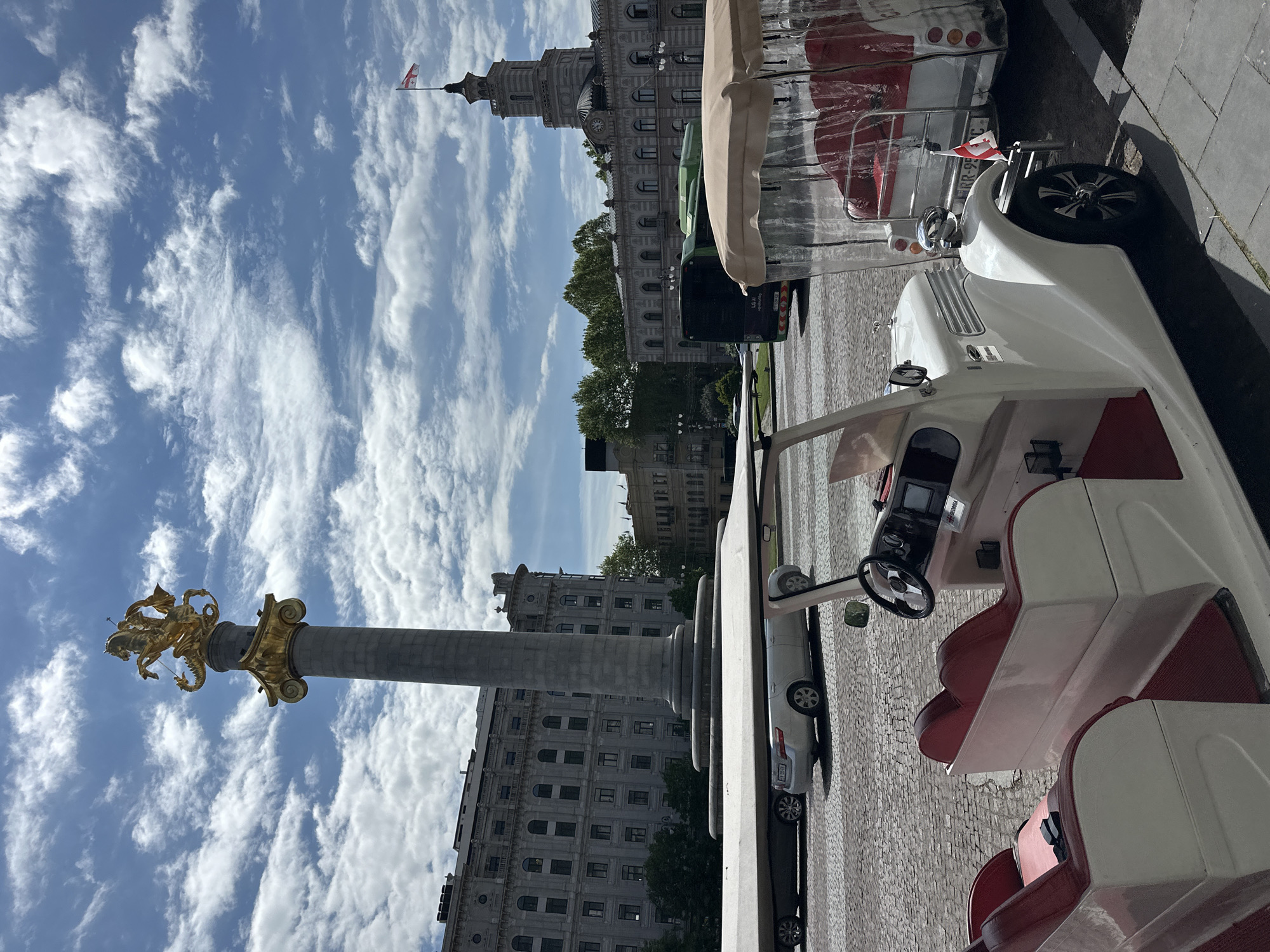 The image depicts a scene in Tbilisi, Georgia, featuring the Freedom Monument and a distinctive white and red tourist vehicle. The monument consists of a tall column with a golden statue of St. George slaying a dragon at its apex. Several older buildings with classical architecture can be seen in the background, partially obscured by trees and the monument. The sky is bright blue with fluffy white clouds. A small Georgian flag is visible on a building in the background. The foreground is dominated by a white tourist vehicle, with red seats, partially visible from the side, parked on a cobblestone street.