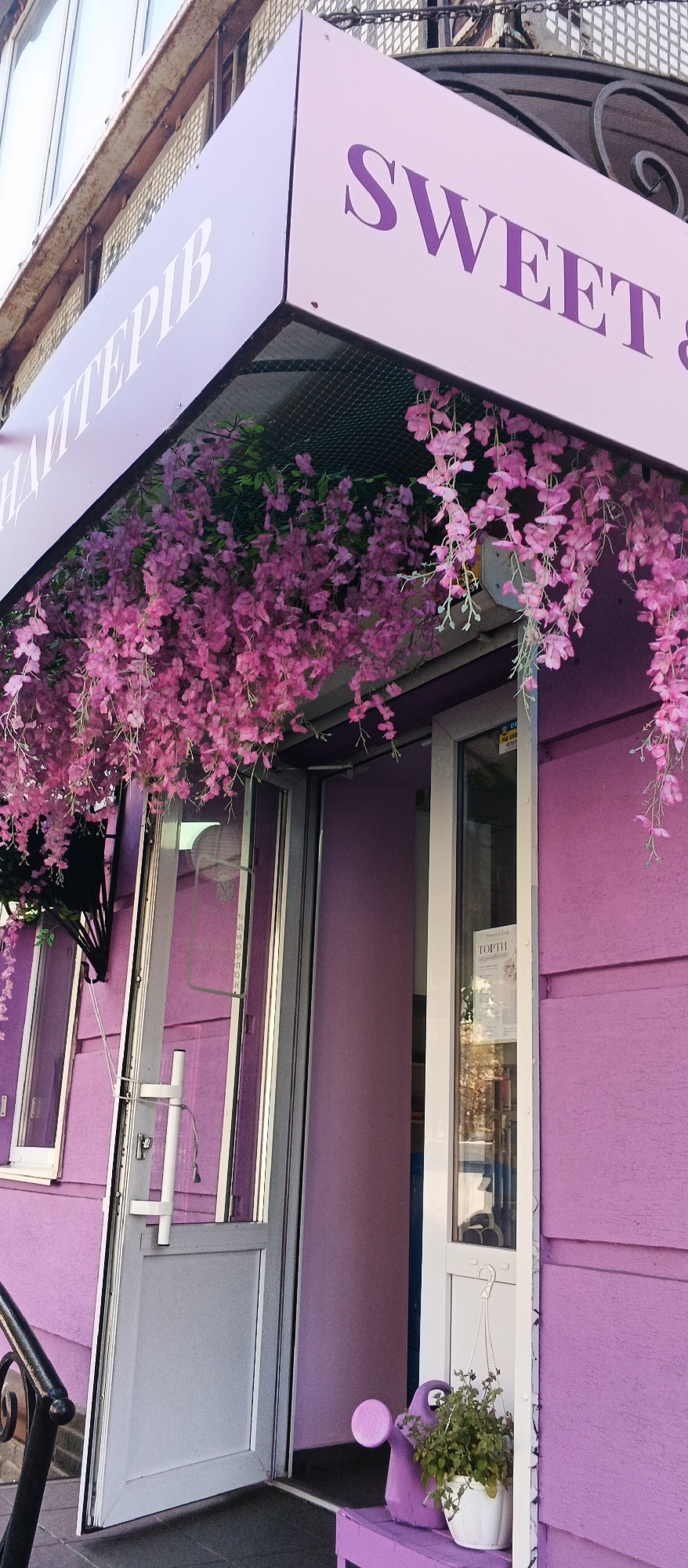 The image shows the exterior of a building with a prominent lilac-colored facade and a sign above the entrance. The sign, also in lilac, features dark purple lettering that reads "SWEET &" on the right side and what appears to be Ukrainian text on the left. Pink artificial wisteria flowers hang from a mesh grid under the sign, obscuring part of the entrance.
The entrance consists of a double door; the left door is gray and slightly ajar, revealing a partial view of the interior. The right door is a glass pane with a white frame, and a white hanging basket with a small plant is suspended from it. A sign inside the glass door displays text, including the word "ТОРТІ" (cakes in Ukrainian) and an illustration.
To the right of the entrance, a small purple bench is visible, with a purple watering can and a white pot containing a green plant on it. The ground in front of the entrance is paved. A black metal railing is partially visible in the lower left corner. The building appears to be located in a commercial or public area, given the storefront-like appearance. The overall aesthetic suggests a bakery or confectionery shop. Based on visual cues and the likely language on the signage, the location can be inferred as Cherkasy, Ukraine.