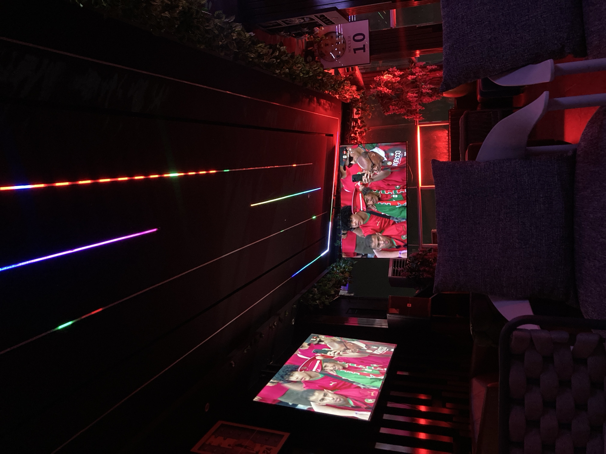 The image shows the interior of a dimly lit sports bar or lounge, likely in Abuja, Nigeria. Two large screens display a soccer match, with players in red and green jerseys. Above the screens, colorful LED strip lights run along the ceiling, casting vibrant hues of purple, blue, green, and red. In the foreground, plush, dark grey seating with intricate weave patterns is visible, suggesting a comfortable viewing experience. A sign with the number "10" and the word "SOCCER" is mounted on a wall to the right, further indicating the sports-themed nature of the establishment. The overall ambiance is modern and atmospheric, designed for patrons to watch sporting events.