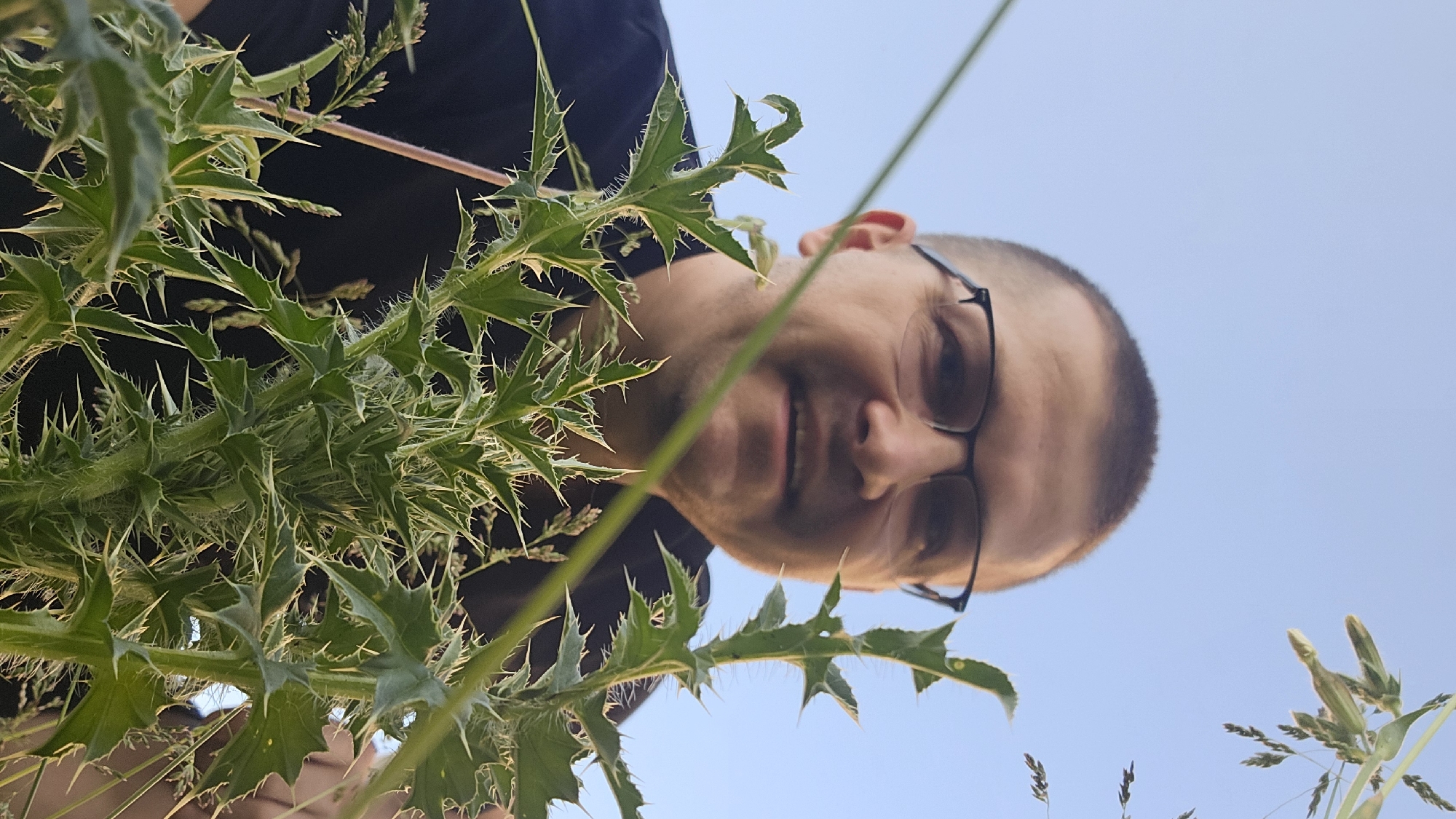 A medium shot, eye-level, outdoors photo shows a man wearing glasses and a black t-shirt looking down at the camera from a low angle, with a clear blue sky in the background. He is partially obscured by prickly green plants in the foreground. The man has short, dark hair and is smiling. The scene appears to be in a natural setting, possibly a park or field, in Kyiv, Ukraine.
