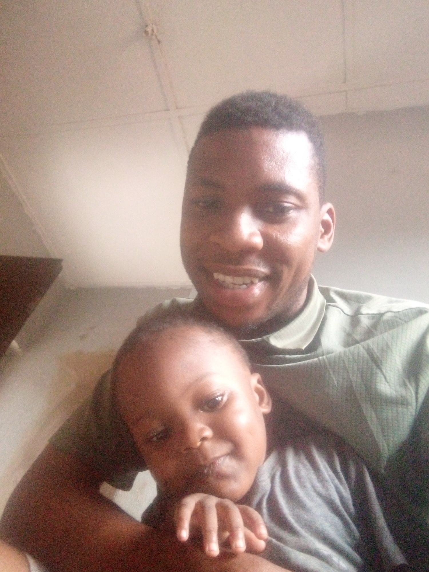 A close-up, eye-level portrait features an adult male and a young child, likely a father and son, sharing a tender moment. Both individuals appear to be of African descent. The adult male, positioned behind the child, is smiling broadly towards the camera, showing his teeth. He wears a textured olive green collared shirt.
The child, held securely in the adult's arms and resting against his chest, looks slightly downwards or to the side with a soft, gentle expression. The child wears a plain grey t-shirt, and their small fingers are visible in the foreground, resting near their chest.
The scene is set indoors, with a simple, light-colored background. The upper left corner reveals part of a ceiling with visible lines, possibly indicating panels or structural elements. The lighting is soft and ambient, suggesting the photograph was taken during daytime. The overall mood of the image is one of warmth, affection, and happiness, particularly evident in the adult's joyful smile. There is no visible text present in the image. The exact location is unknown, but it appears to be a casual indoor setting, possibly a home.