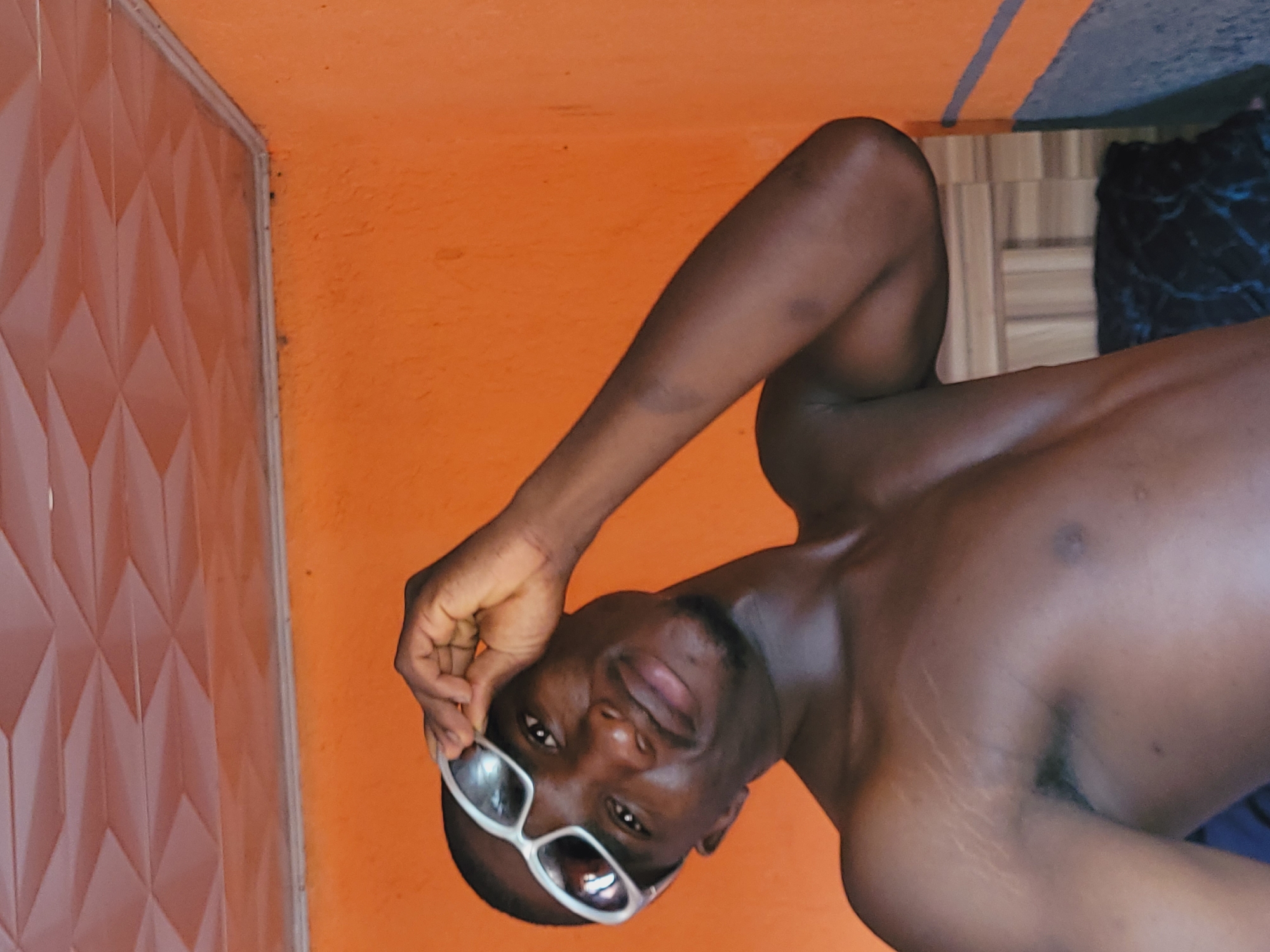 The image displays a dark-skinned man indoors, photographed from a slight low-angle perspective. The image is rotated 90 degrees counter-clockwise; if viewed upright, the man would be standing or sitting, looking slightly upwards towards the camera.
The man is shirtless, revealing his upper torso and arms. His left arm is bent, with his hand near his head, holding a pair of white-framed sunglasses that are resting on his face. He looks directly at the viewer with a neutral, calm expression. Notable details include visible stretch marks on his left shoulder and upper arm.
The background primarily features a vibrant orange wall. To his left (or the top of the image in its current orientation) is a section of a textured wall or door, characterized by a repeating geometric, diamond-like pattern in a brownish-pink shade. A thin, light-colored frame separates this patterned surface from the orange wall. In the bottom right corner, indistinct elements like dark, patterned fabric and a wooden surface are partially visible. The scene suggests an interior setting, likely within a building in Bukuru, Nigeria. No text is visible in the image.
