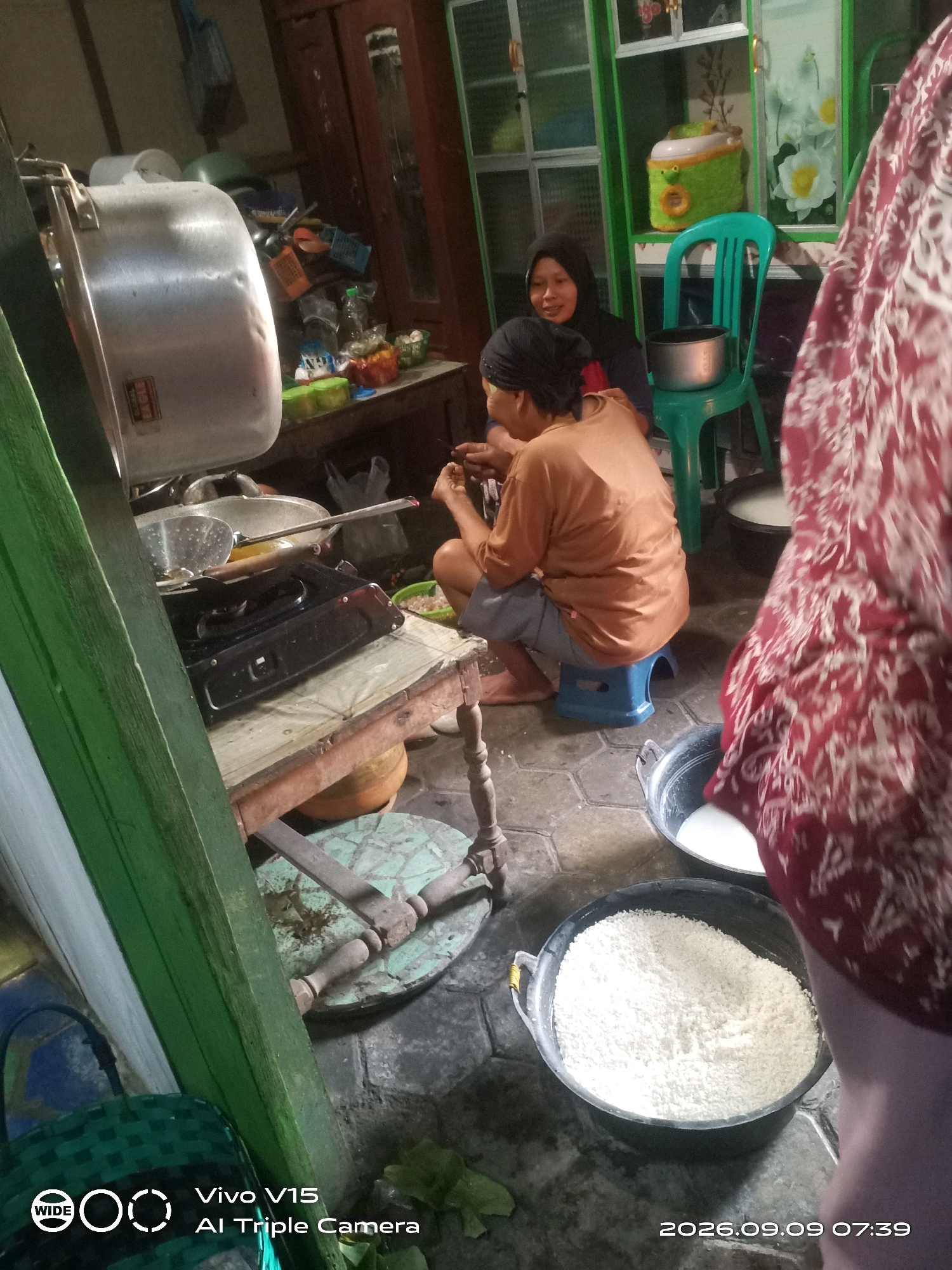 In an indoor, low-light scene, two women are present in what appears to be a kitchen area. The woman on the left, wearing a black hijab, is seated and looking towards the right. The woman on the right, wearing a brown shirt and blue shorts, is also seated and is engaged in an activity, possibly preparing food, as her hands are occupied.
The environment is cluttered with various objects. To the left, a large metallic pot is prominent, suggesting cooking activities. A black gas stove is situated on a wooden table, with cooking utensils nearby. Several plastic containers and bags are visible on shelves and surfaces, along with a green chair. In the foreground, two large black basins are present; one contains a white granular substance, possibly flour or rice, and the other appears to contain a white liquid. The floor is tiled with a hexagonal pattern. A large red and white patterned fabric, possibly a garment, is draped in the foreground, obscuring part of the scene.
The image has a timestamp overlay indicating "2026.09.09 07:39" and "Vivo V15 AI Triple Camera", which is likely metadata from the device used to capture the photograph. The location is identified as "Plosorejo, Indonesia".