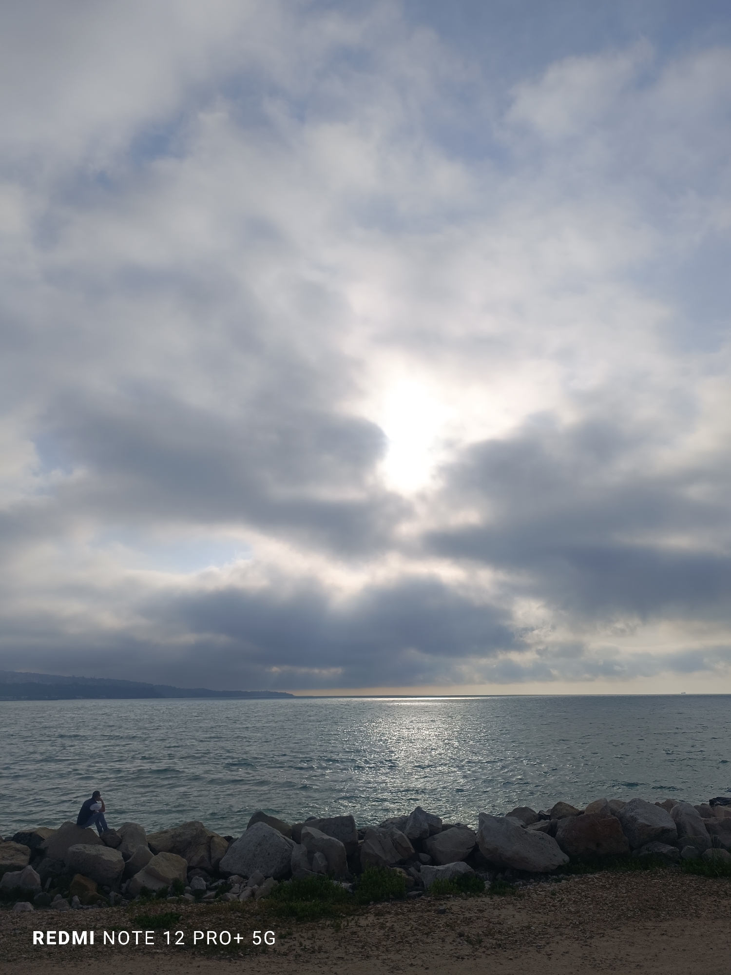 This outdoor photograph captures a contemplative coastal scene, likely along the Black Sea in Varna, Bulgaria. The main subject is a single individual, dressed in dark clothing, sitting on a large rock in the foreground. The person faces away from the viewer, looking out across the vast expanse of water.
The setting features a rugged rocky shore in the immediate foreground, with scattered green vegetation growing between the stones and on the sandy ground. Beyond the rocks, the sea stretches out to the horizon, its surface reflecting the bright light. In the far distance on the left, a faint landmass or coastline is visible, appearing hazy.
The weather is cloudy, with heavy grey clouds dominating the sky. However, the sun dramatically breaks through a central clearing in the cloud cover, casting brilliant rays and creating a strong, shimmering reflection on the water. This suggests a time of day when the sun is relatively low, either morning or late afternoon, providing a diffused yet powerful light.
The overall mood conveyed by the lone figure and the expansive natural elements is one of quiet solitude or reflection.
In the bottom left corner of the image, white text is visible, stating "REDMI NOTE 12 PRO+ 5G," indicating the device used to take the photograph.