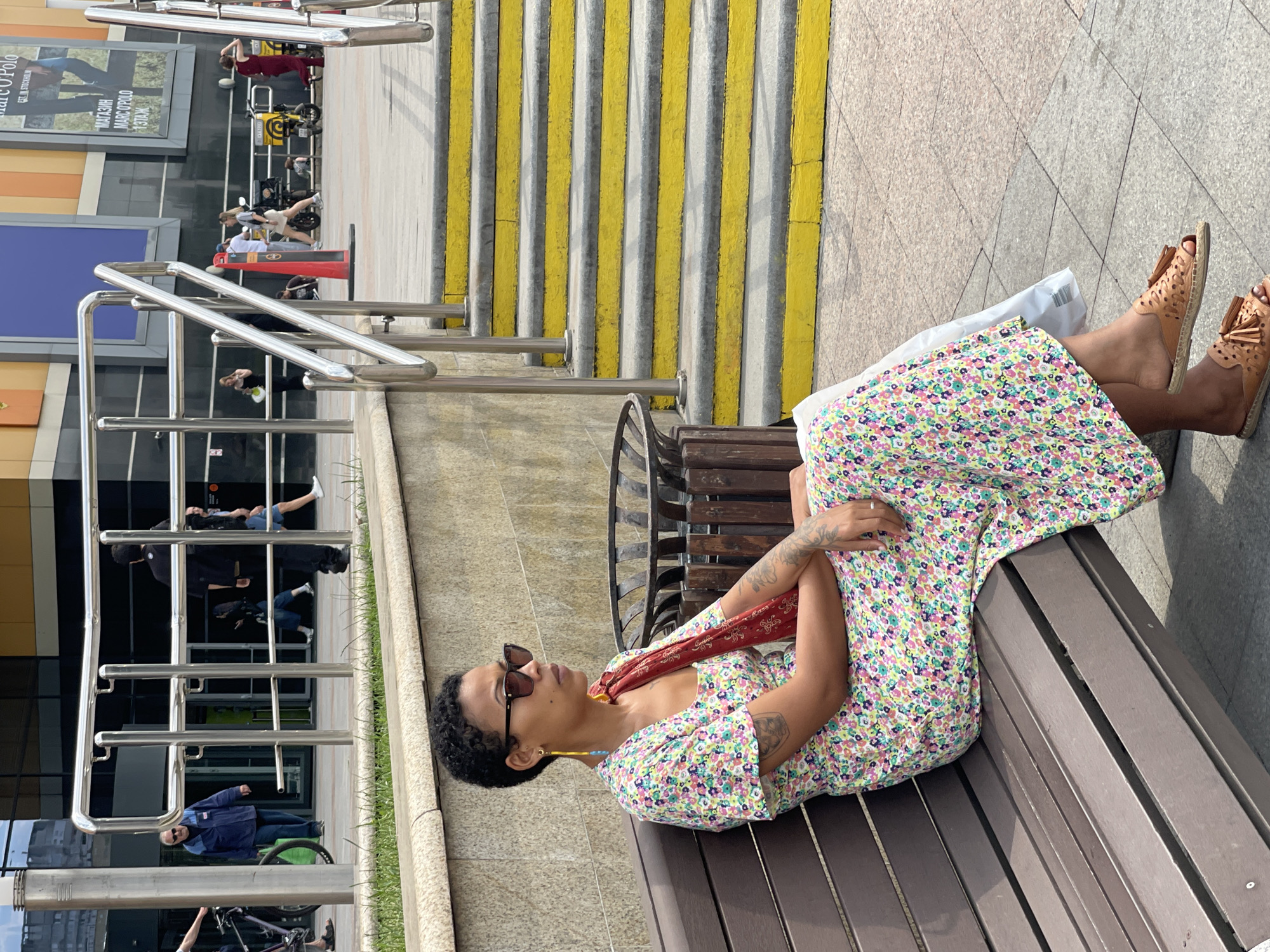 The image depicts a woman with short, dark hair and sunglasses sitting on a wooden bench. She is wearing a floral-print dress and tan sandals. A red scarf is draped over her shoulder. In the background, there are stairs with yellow treads and handrails. Several people are walking in the distance, and there is a building with large windows. The scene appears to be outdoors in a public area, likely a pedestrian walkway or plaza. The overall impression is a casual daytime scene in an urban environment. The presence of Cyrillic text on a sign in the background indicates the location is Moscow, Russia, specifically near a shopping center given the poster advertising "MARC O'POLO".