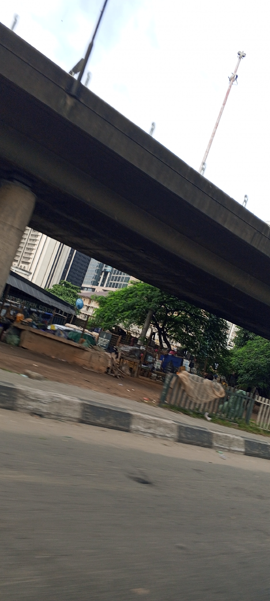 The image shows a street scene in Lagos, Nigeria, viewed from a moving vehicle. In the foreground, a blurred asphalt road is visible with some potholes. A concrete curb with black and white markings lines the side of the road. Beyond the curb is a dirt area with various stalls and structures, some made of corrugated metal and wood, with tarpaulins stretched over them. Inhabitants are partially visible behind these structures, with some appearing to be vendors. Trees with dense green foliage are interspersed among the stalls and buildings. In the background, modern high-rise buildings with glass facades stand tall, contrasting with the makeshift structures at street level. Overhead, a large concrete flyover or bridge dominates the upper portion of the image, obscuring a portion of the sky. A tall, thin metallic structure, possibly a crane or antenna, is visible against the overcast sky. The overall impression is a busy urban environment with a mix of infrastructure and informal commerce.