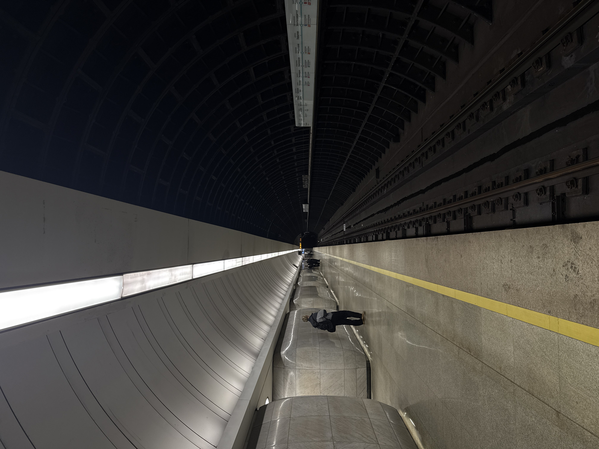 The image depicts the interior of a subway station, likely in Moscow, Russia, based on the architectural style. The station features a high, arched ceiling with a grid-like pattern. Along one side, there's a curved wall illuminated by long, rectangular light panels. The opposite side shows the tracks. A yellow safety line runs along the edge of the platform. Several people are waiting on the platform. The perspective suggests the photo was taken from a high vantage point.