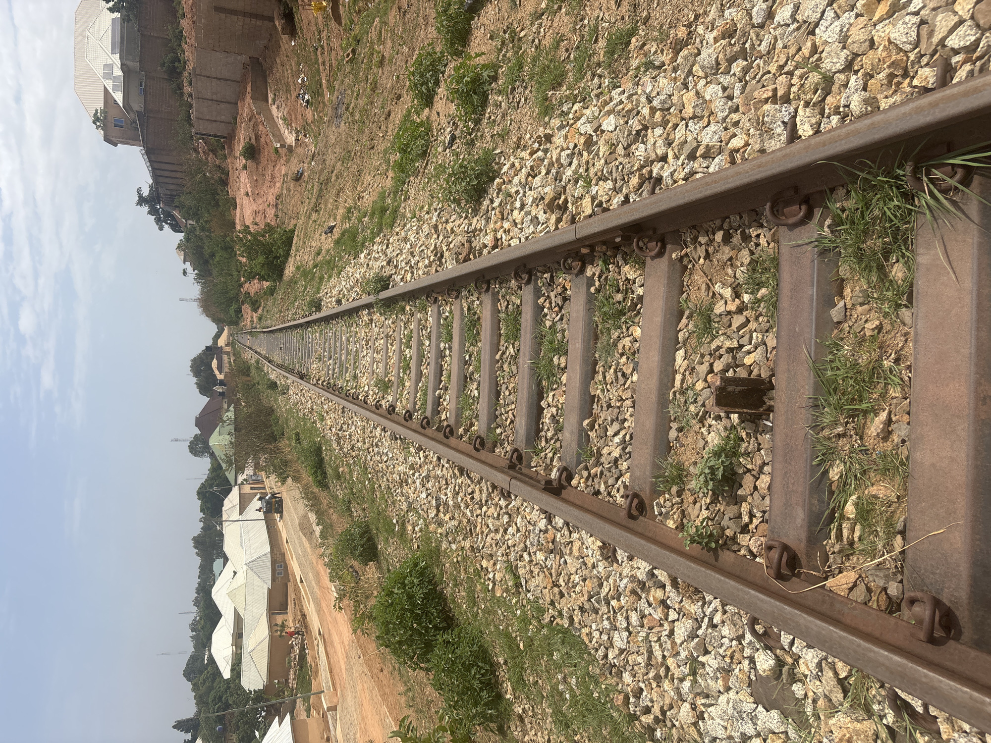 The image depicts a railway track running through an urban area in Jos, Nigeria. The tracks are constructed with weathered steel rails secured to concrete sleepers, surrounded by gravel ballast. Vegetation, including grass and small plants, grows between the sleepers and alongside the tracks. In the background, there are buildings, including residential houses with corrugated metal roofs, and a retaining wall structure with a building on top of the upper ground level. The sky is overcast, suggesting a hazy or cloudy day.