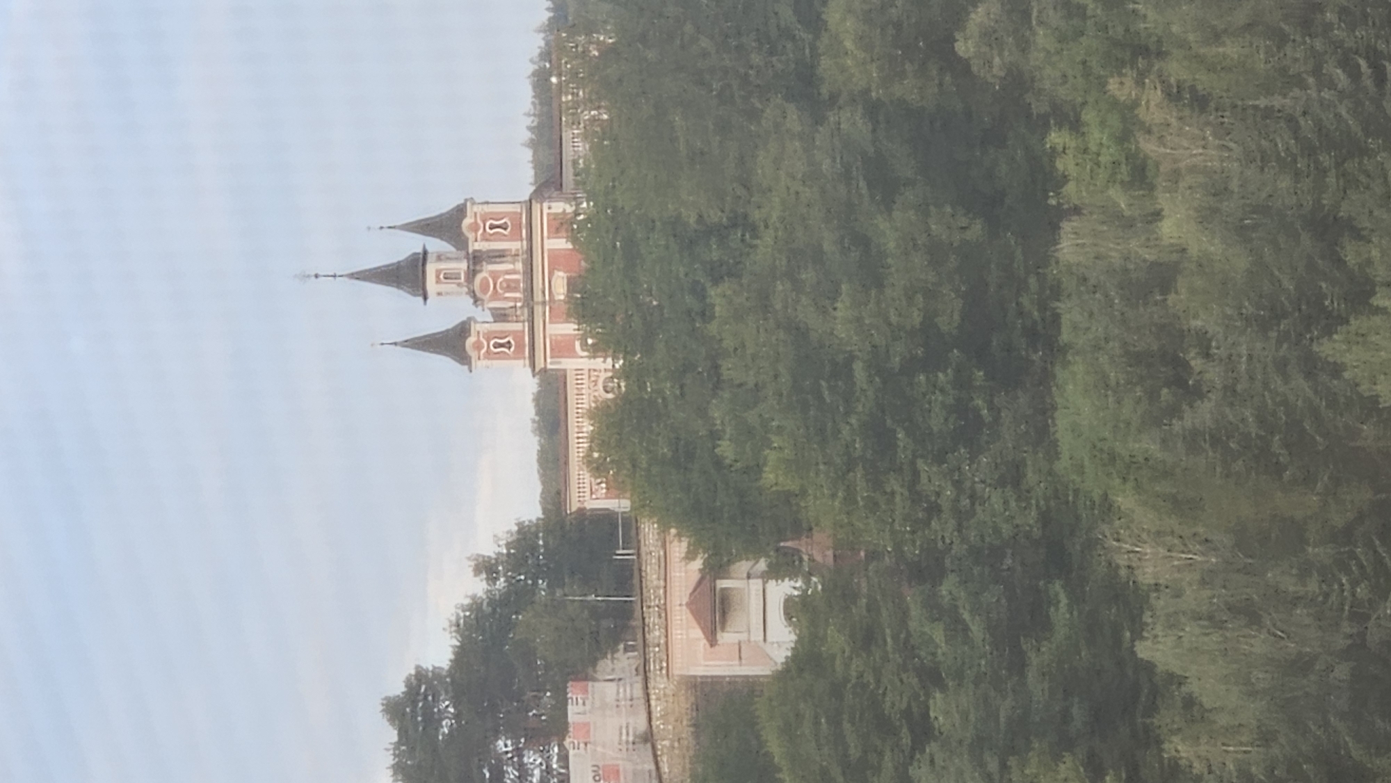 The image depicts a view of the Holy Trinity Church in Prešov, Slovakia, a striking building characterized by its distinctive red and white facade and three prominent spires. The church is situated atop a hill, surrounded by lush green trees that partially obscure the lower portions of the structure. In the foreground, dense foliage creates a natural barrier, emphasizing the church's elevated position. The sky above is a pale, hazy blue, suggesting either an overcast day or early morning light. To the left of the church, construction scaffolding is visible, indicating ongoing renovation or building work. A small, light-pink building with a small tower is nestled among the trees below the main church structure. The overall scene is one of architectural beauty set within a verdant, natural landscape.
