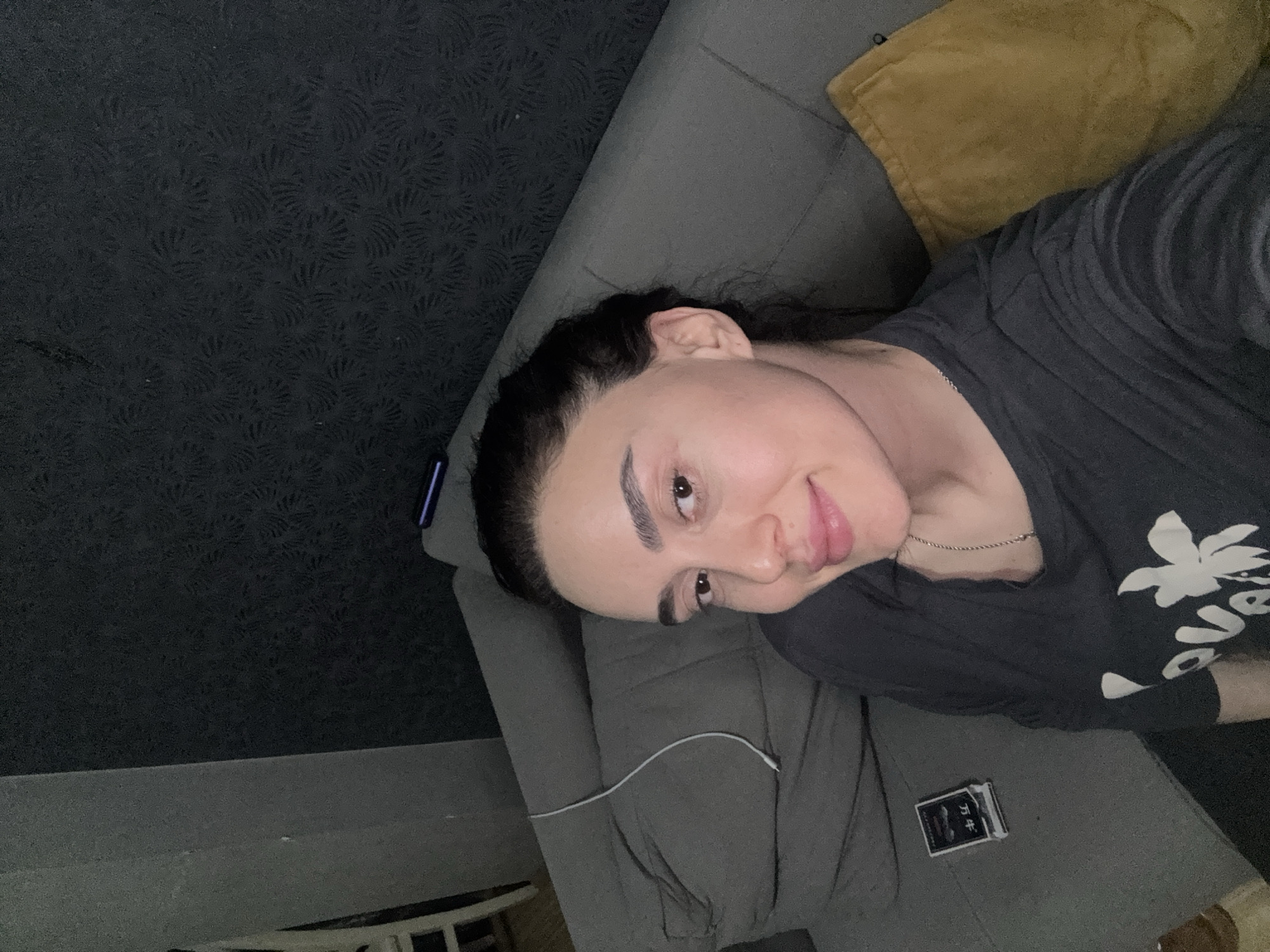 The image is a selfie of a woman lying on a grey couch. She is wearing a dark t-shirt with a white design. A yellow pillow is visible behind her. A white charging cable is resting on the couch near her. A small, black book or package with text in a foreign language is placed next to her. The wall behind the couch has a dark, patterned wallpaper. The location is not discernible from the image; there is no indication it is in Tbilisi, Georgia.