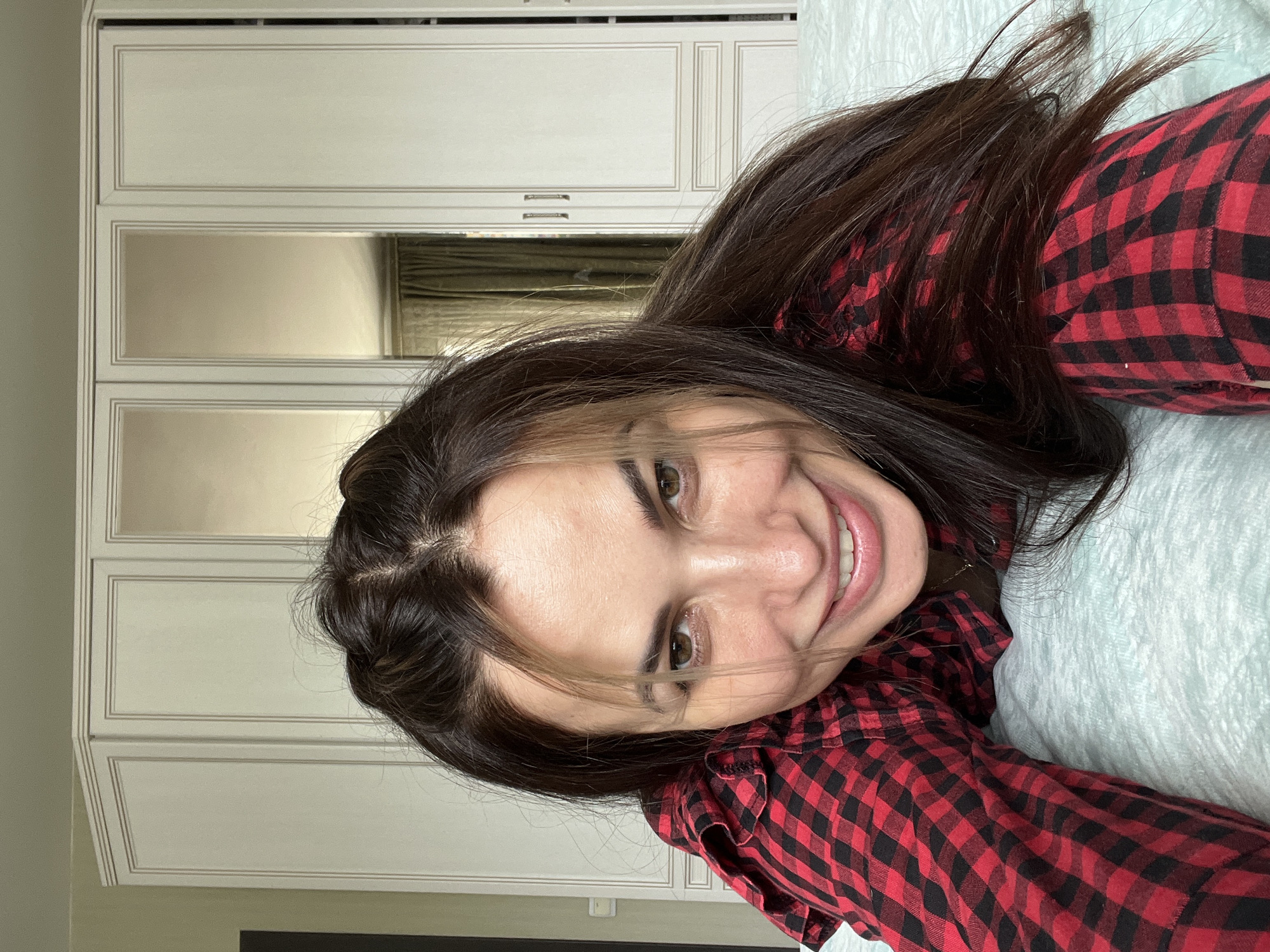 A young woman with long, dark brown hair is captured in a close-up portrait, smiling broadly at the camera. She is wearing a red and black plaid shirt, with her head tilted, making the image appear rotated. Her face shows a joyful expression, with her light brown or hazel eyes wide and her lips parted in a wide smile, revealing her teeth. strands of hair frame her face and fall across her forehead.
She appears to be lying down, with her body mostly out of frame, but her upper torso and a portion of her plaid shirt are visible. A light blue or greenish textured fabric, possibly a blanket or bed sheet, is visible beneath her and to the right.
The background consists of light-colored, possibly white or off-white, wooden furniture, which appears to be a wardrobe or built-in cabinets. The furniture has decorative moldings and includes mirrored panels in its middle section. These mirrors reflect what looks like a green or teal curtain or blinds and other ambient room elements. The setting is clearly indoors. There are no visible texts or specific time-of-day cues. The overall mood of the image is cheerful and casual.