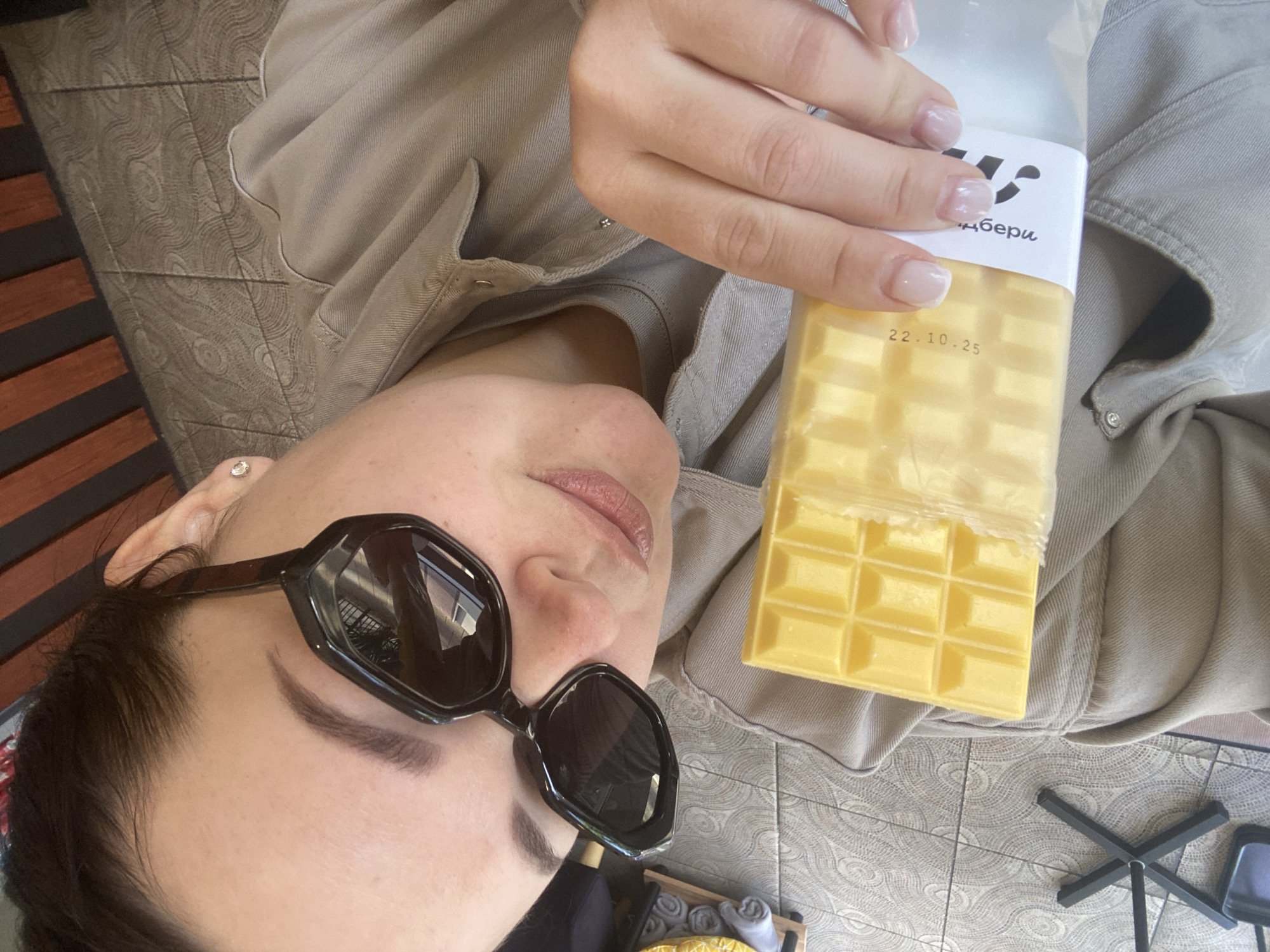An overhead, slightly angled shot captures a person, likely a woman, lying down and holding a package of yellow, segmented bars. The person is wearing a light beige or khaki-colored jacket or shirt, and has dark hair, a subtle pink lip color, and natural-looking eyebrows. She is wearing dark, hexagonal-framed sunglasses, which reflect a bright, sunny outdoor or semi-outdoor scene. A small stud earring is visible in her right earlobe. Her left hand, adorned with a French manicure, holds the clear plastic package.
The package contains a block of bright yellow, rectangular segments, resembling a chocolate bar or perhaps wax melts. A white label on the package features part of a Cyrillic word, "берu," which could be part of a brand name like "Wildberries" (Валдберри), a common online retailer in Russia. Below this, the date "22.10.25" is printed, likely indicating an expiration or production date.
The background shows a patterned tiled floor with concentric circle designs, suggesting a patio or similar outdoor setting in Sochi, Russia. A dark, possibly metal, stand or furniture leg is visible in the lower right, and a wooden slatted structure can be seen on the far left. The overall impression is one of relaxation, perhaps enjoying a snack or product outdoors on a bright day.