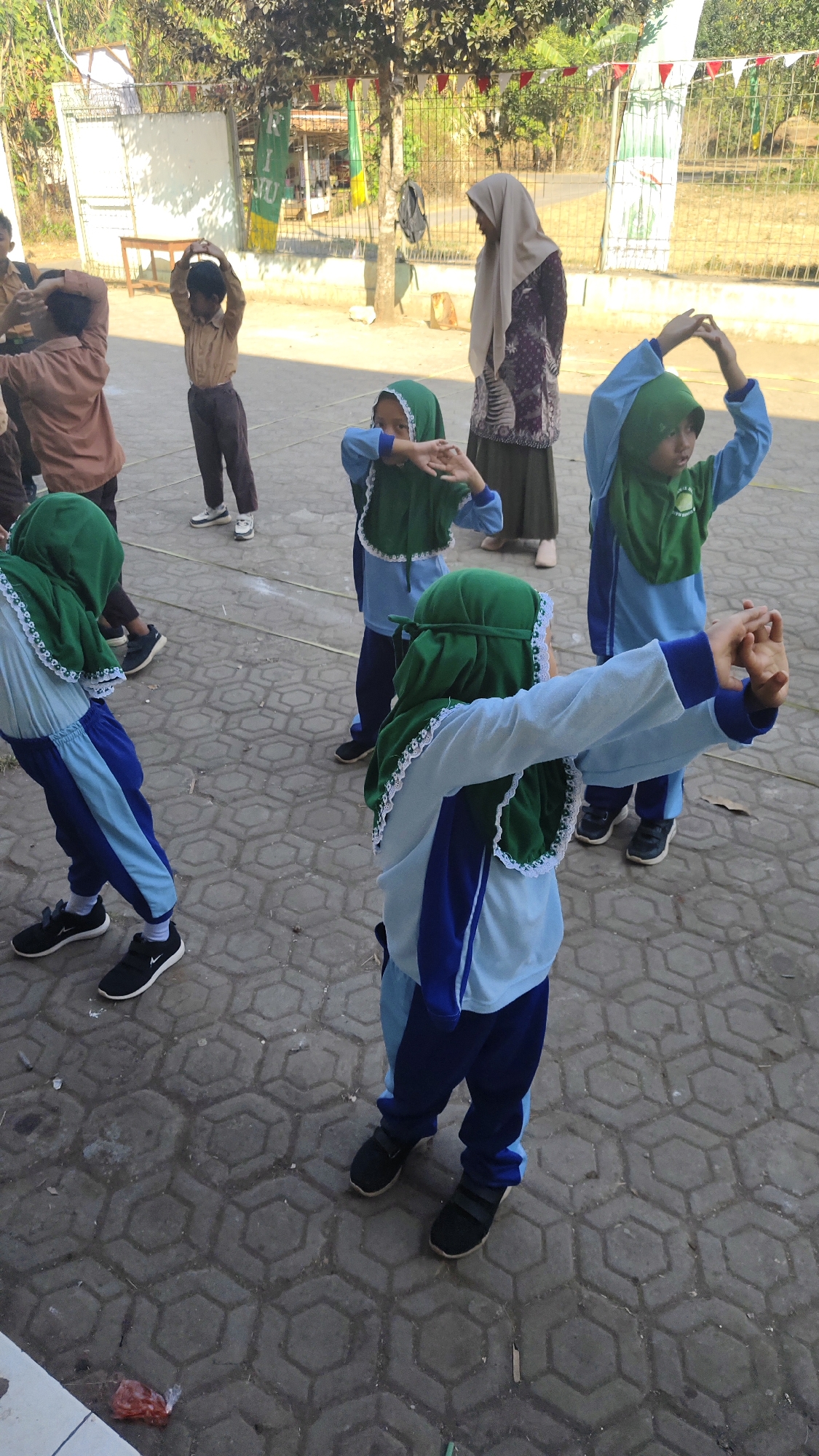 A group of children, likely students, are engaged in physical activity outdoors on a paved courtyard. They are wearing uniforms that include light blue shirts and dark blue pants, along with green hijabs. Several children have their arms raised and extended, suggesting they are performing exercises or a dance. One adult woman, wearing a hijab and a patterned dress, stands in the background, observing the children. The environment appears to be a school or community area, with trees and fencing visible behind the group. The pavement is made of hexagonal concrete blocks. The scene is illuminated by sunlight, casting shadows on the ground. The image is taken from a slightly elevated, eye-level perspective.