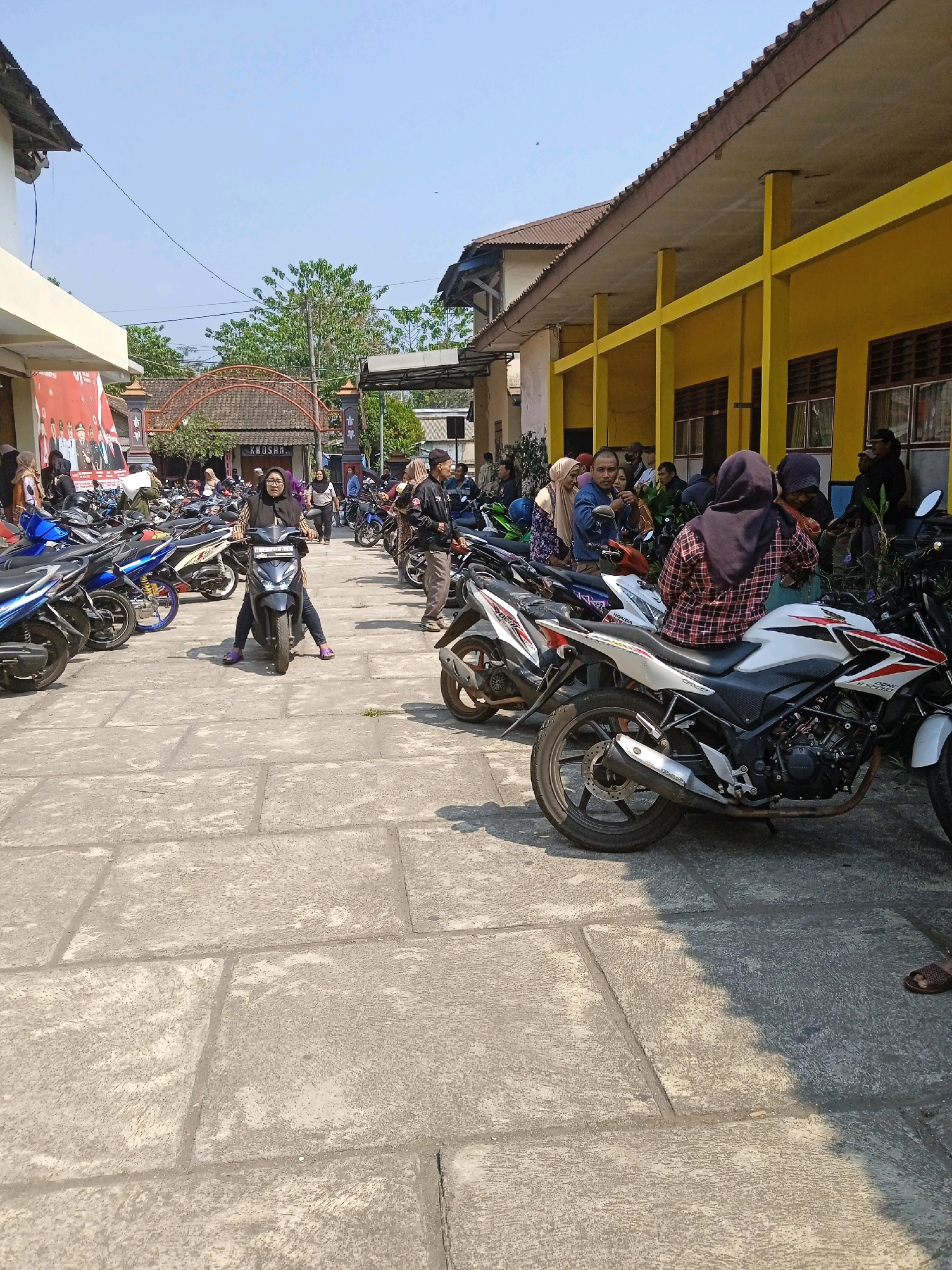 This image depicts a sunny day scene in a courtyard or parking area outside a yellow building, likely a public institution or school, located in Bedono, Indonesia. The ground is paved with large, light-colored rectangular stone tiles. A multitude of motorcycles are parked in rows, filling a significant portion of the foreground and midground.
In the foreground, a woman wearing a dark hijab and a patterned red and black shirt is seated on a white and red motorcycle. Her feet are visible, wearing light-colored shoes. Behind her, more motorcycles are parked, with their handlebars and seats visible.
In the midground, a woman in a dark hijab is sitting on a dark-colored scooter, looking towards the camera. Several other people, some wearing hijabs, are gathered around or walking amidst the motorcycles. A large banner with portraits of uniformed individuals is visible on the left side of the image, attached to a building.
The background reveals more buildings, including a yellow structure with dark-framed windows and a brown tiled roof. A red-arched gateway with decorative elements is also visible, with the word "KHOSAR" inscribed on it, possibly indicating a name or location. Lush green trees can be seen behind the gateway. The sky is clear and bright blue, indicating fair weather. The overall impression is a busy public space with a significant number of motorcycles, suggesting a gathering or event.