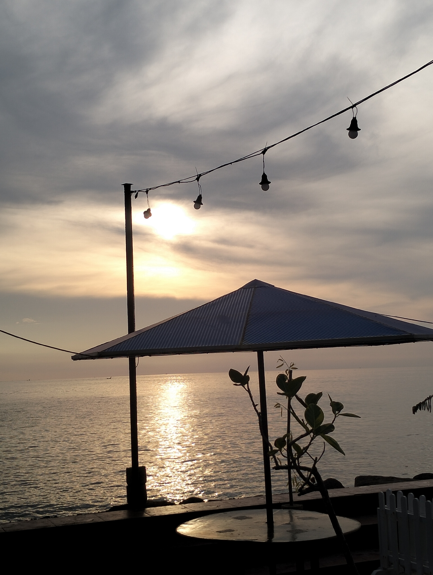 The image depicts a coastal scene at dusk, likely in Lhokseumawe, Indonesia. The sun is setting, casting a golden reflection on the sea. A string of lights is suspended overhead, with bare bulbs hanging from the wire. A blue, pyramid-shaped awning covers a table. A small plant with sparse leaves stands beside the table. A low wall borders the scene, with a white picket fence visible on the right. The overall scene is serene and dimly lit, silhouetted by the setting sun. A distant boat can be seen on the water.
