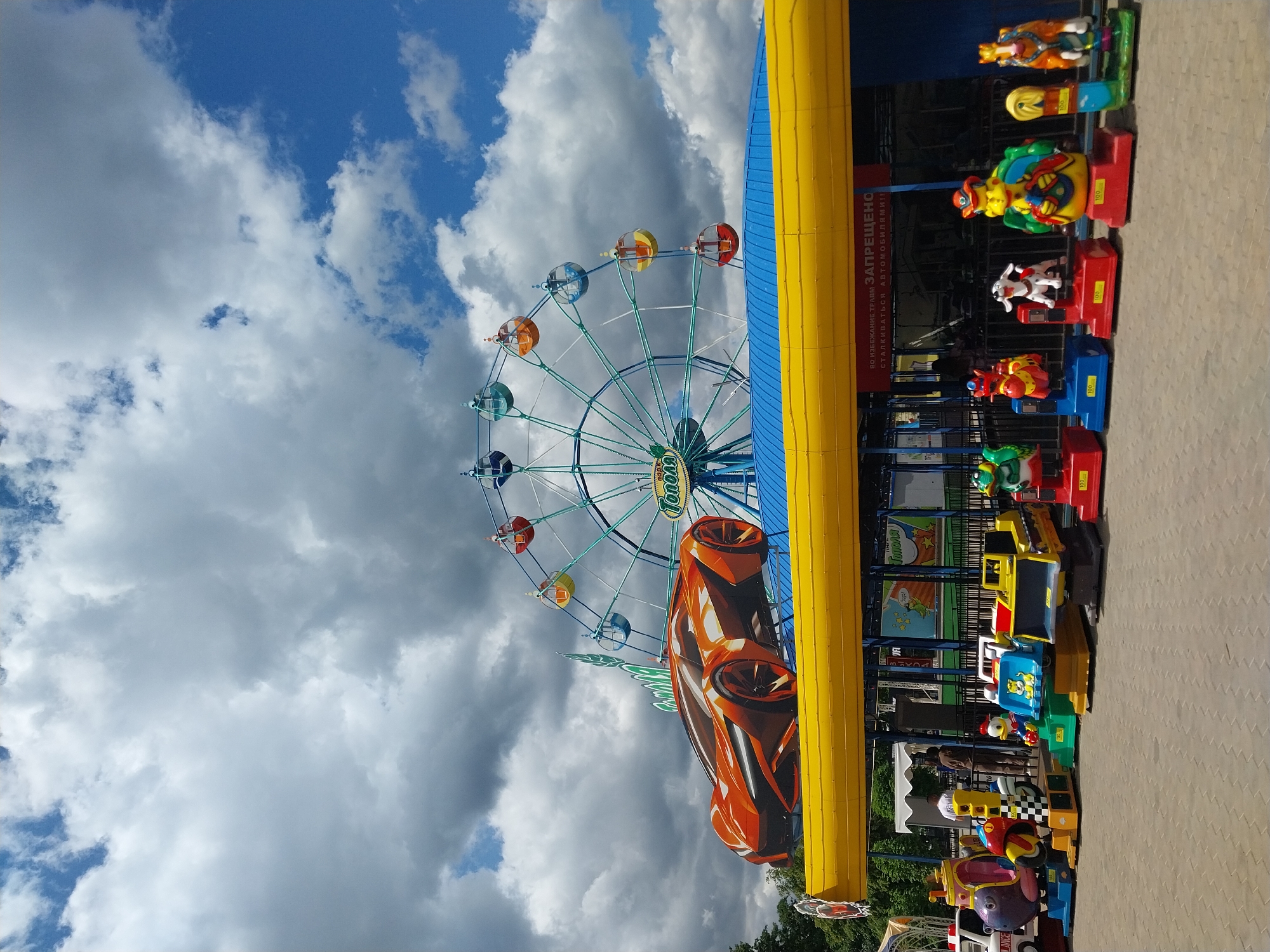 The image depicts a vibrant scene at a children's amusement park in Orenburg, Russia. The amusement park features a Ferris wheel and a row of coin-operated rides for children. The rides are themed after various animals and vehicles, including a race car, a police car, and cartoon characters. The sky above is partly cloudy with both blue sky and large, puffy clouds. The scene appears to be captured during daylight hours, with the sun illuminating the area. A large, yellow structure is adjacent to the children's rides. There are people visible standing behind the row of children's rides.