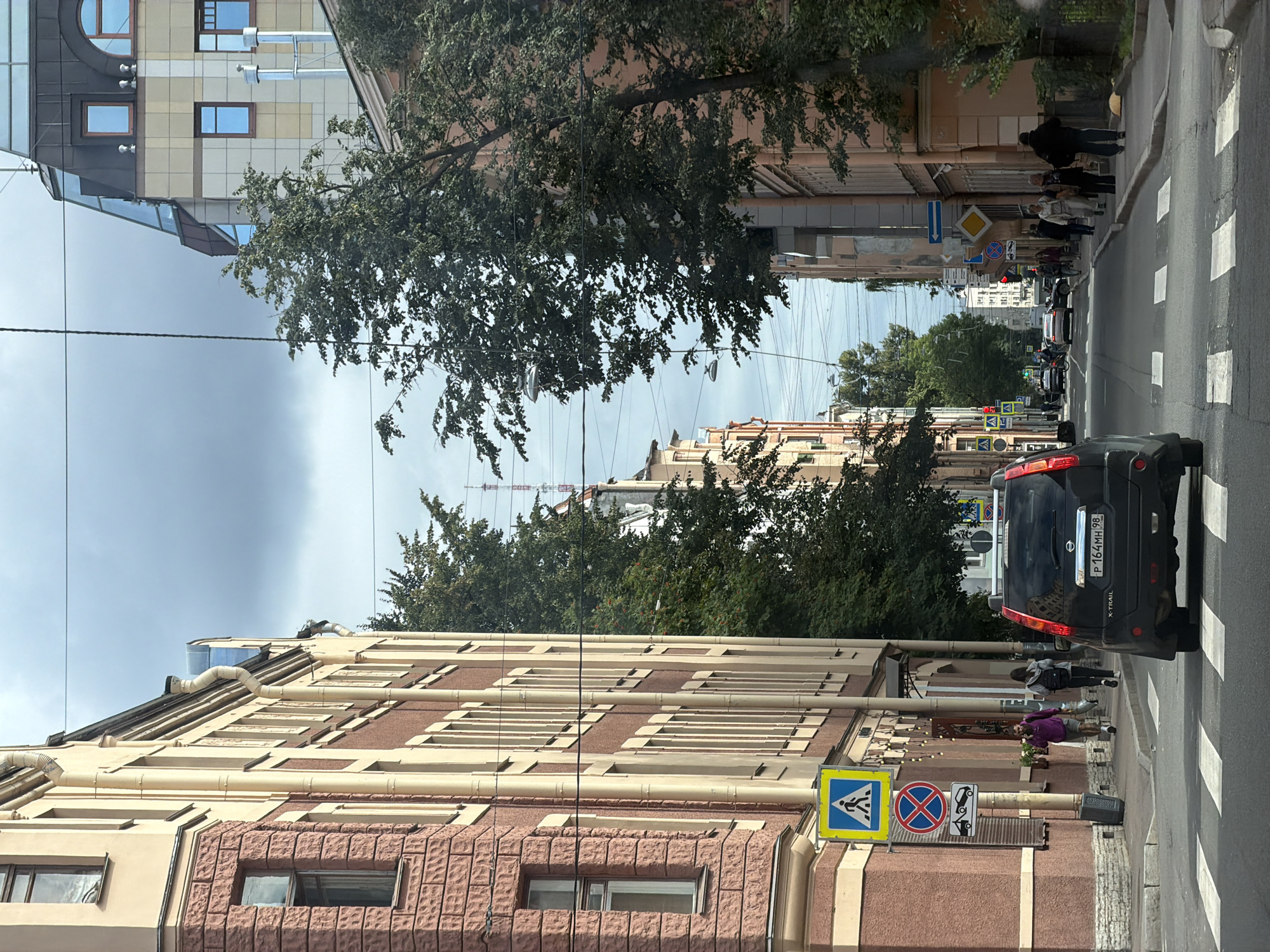 The image depicts a street scene in Saint Petersburg, Russia, likely on a cloudy day, as indicated by the overcast sky. The perspective is from a moving vehicle, facing forward down a paved street lined with multi-story buildings.
On the left, a building with a reddish-brown stone lower facade and a lighter beige upper facade features several windows. A yellow pedestrian crossing sign with a blue border and a white pedestrian symbol is visible on a pole on the sidewalk. Below it, there's a circular red sign with a diagonal line and a tow truck symbol, indicating no parking or towing. The sidewalk has a few pedestrians.
On the right, the buildings are also multi-story, with a more modern building at the far end featuring a glass-paneled section on its roof. Trees with green foliage are present along both sides of the street, partially obscuring the buildings and creating a canopy effect in some areas. Overhead, numerous electrical wires crisscross the sky, supported by poles.
A black Nissan X-TRAIL SUV is in the foreground, partially obscuring the right side of the image. Its license plate is visible as "P 164MH 98". The vehicle is positioned over a pedestrian crossing marked with white stripes on the asphalt. Further down the street, other vehicles, including a white sedan, are visible, along with more pedestrians on the right sidewalk. Traffic lights and street lamps are also present. The overall impression is a typical urban streetscape in Saint Petersburg.