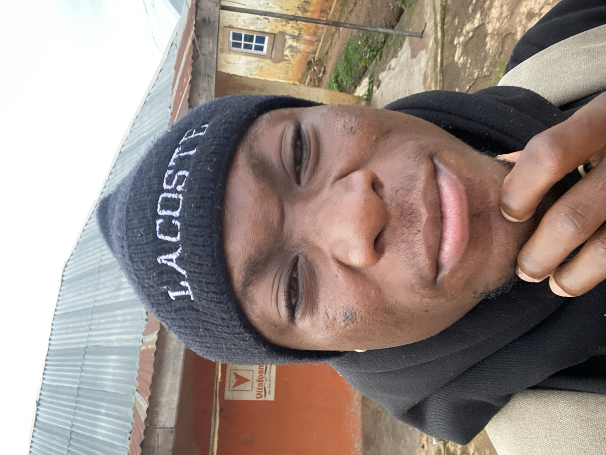 A close-up, selfie-style photograph captures a young Black male wearing a black beanie with white lettering that reads "LACOSTE". He is looking directly at the camera with a contemplative expression, his chin resting on his hand. His lips are slightly parted, revealing a hint of teeth. The background suggests an outdoor setting in Vom, Nigeria, with a corrugated metal roof visible above a building. The building itself has an orange-colored section with a "Vitafoam" logo, and a light-colored wall with a small, multi-paned window. The ground appears to be dirt and concrete. The overall impression is a candid shot taken in a rural or semi-rural environment.