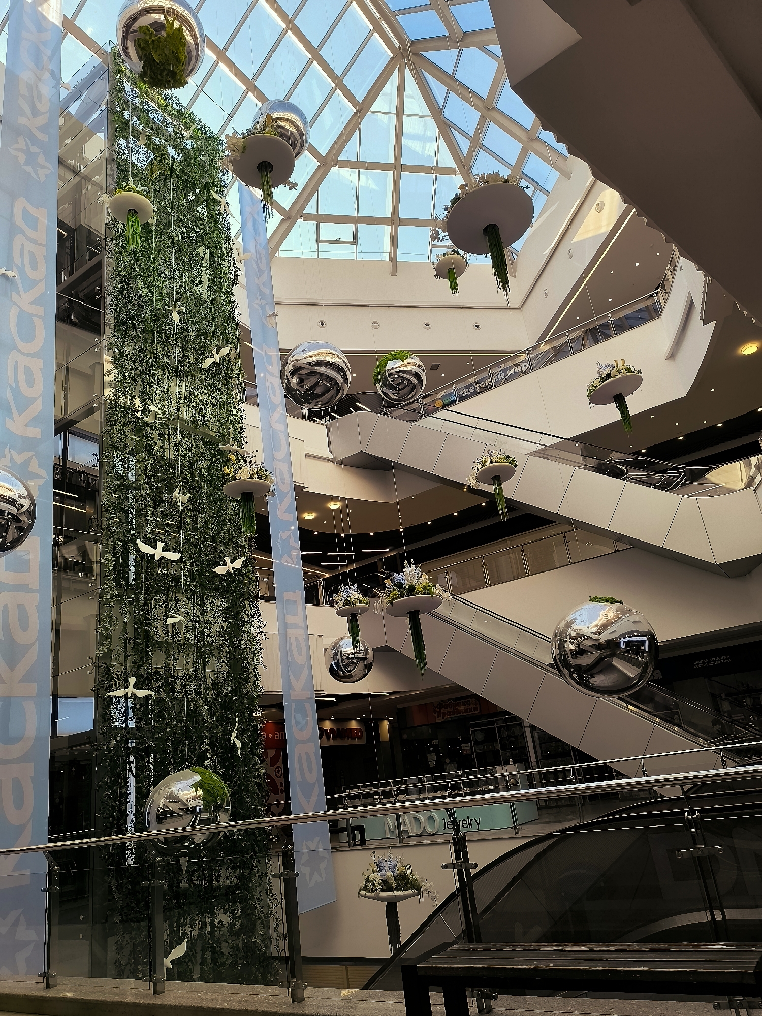 The image depicts the interior of a multi-level shopping mall, characterized by its modern architecture, extensive use of glass, and escalators. A prominent feature is a tall, vertical installation of greenery, resembling a living wall or a large plant column, suspended from the ceiling. Scattered around this green column and throughout the mall's levels are large, reflective spherical ornaments, possibly made of polished metal, which mirror the surrounding environment.
Dangling from the ceiling and at various heights are disc-shaped decorative elements, some of which hold floral arrangements. These hang by thin wires or cords. White, bird-shaped ornaments are also suspended at different levels, appearing to "fly" around the central green installation.
The mall's interior features multiple escalators, glass railings, and various storefronts visible on different levels. The ceiling is a large, geometric skylight, allowing natural light to illuminate the space. Some banners with text, including what appears to be the word "kackan" in Cyrillic script, are visible on the left side of the image. Another banner with the text "MADO jewelry" is visible on a lower level.
The overall impression is that of a well-lit, spacious, and decoratively appointed shopping center, likely designed to create a visually engaging and perhaps nature-inspired atmosphere within an urban environment. The specific location in Qarağandı city administration, Kazakhstan, is not visually evident from the image itself; this information is provided as context for the scene.