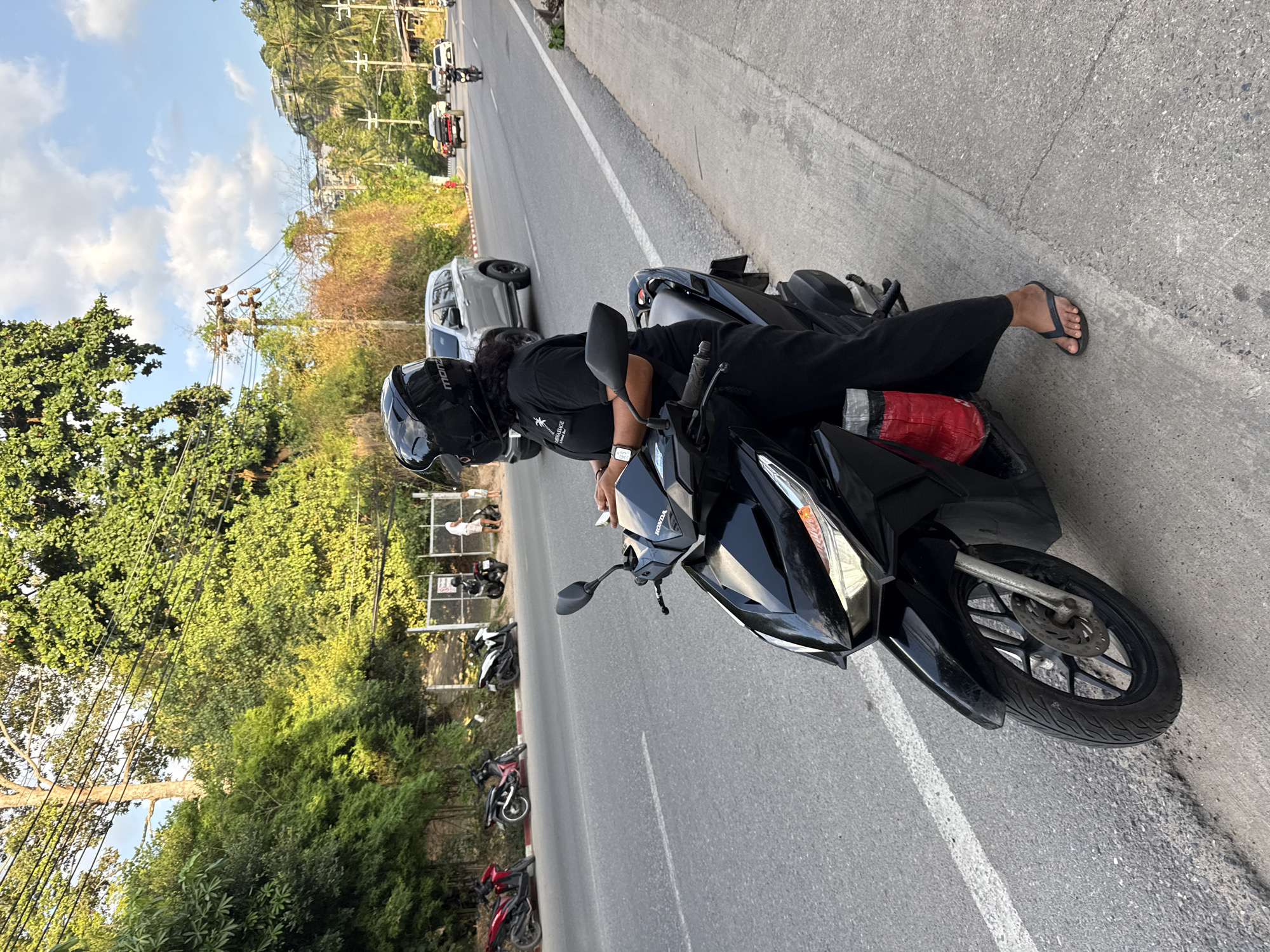 A person wearing a black helmet and black clothing sits on a black Honda scooter on the side of a road in Ko Samui District, Thailand. The person is looking down at a mobile phone held in their hands. A red object is visible tucked into the scooter's storage compartment. The road is paved and has white lane markings. In the background, there are trees, power lines, several parked motorcycles, and vehicles on the road, including a silver SUV and a pickup truck. Buildings are visible on a hill in the distance. The sky is blue with some clouds. The lighting suggests it is either morning or late afternoon.
