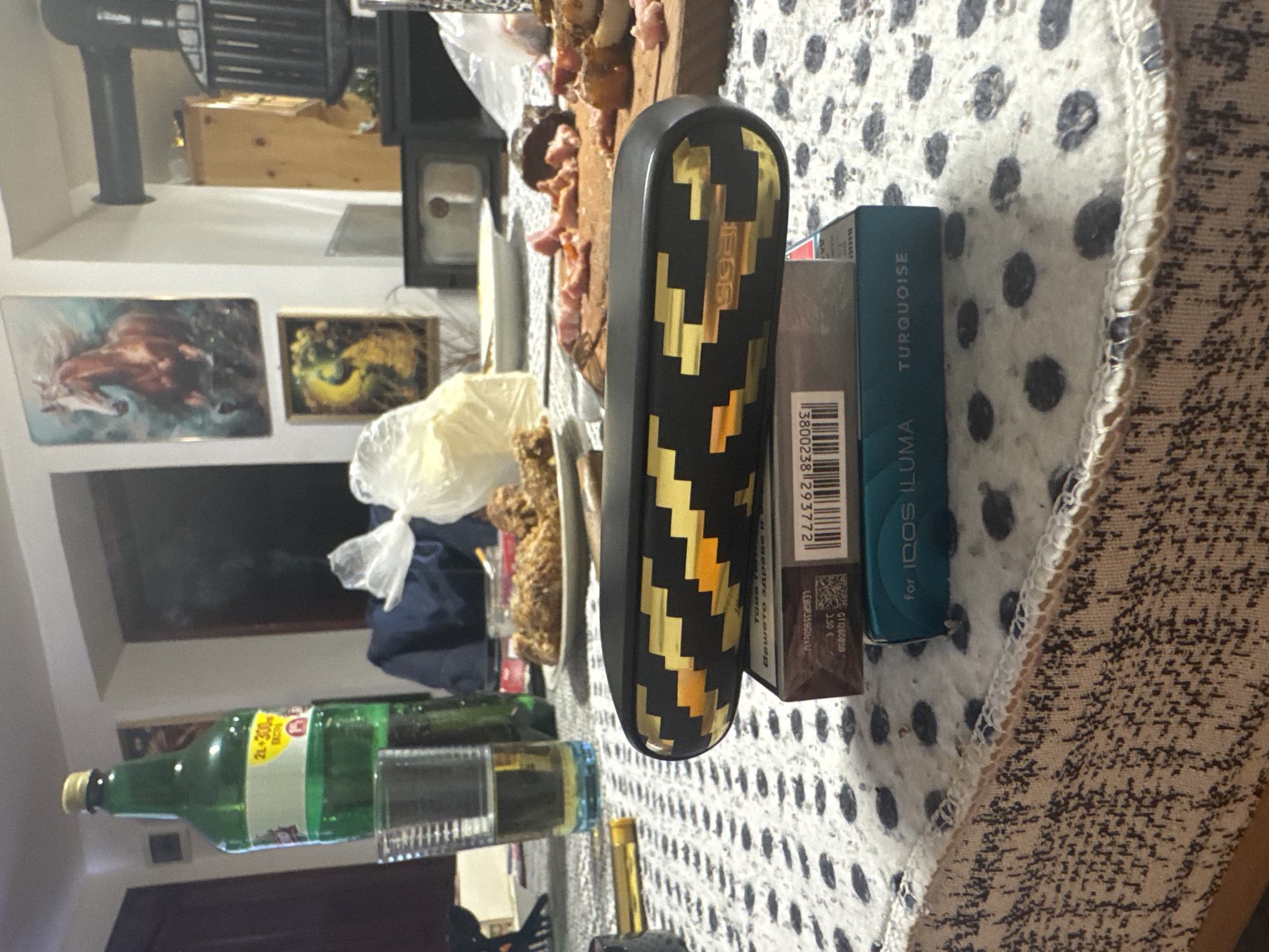 The image captures a cluttered table setting. An Iqos Iluma device with a black and gold pattern is centrally positioned, alongside a pack of Iqos Iluma Turquoise heatsticks. A partially obscured pack of brown cigarettes is also present. A green bottle, possibly of an alcoholic beverage, lies on its side towards the bottom left. Above the table, a glimpse of domestic interior is visible, including wall-mounted artwork and various kitchen appliances. Considering the presence of Bulgarian text on the cigarette packaging, the location is presumed to be in Bulgaria. There is no information to connect the image to the city of Livada in Bulgaria.
