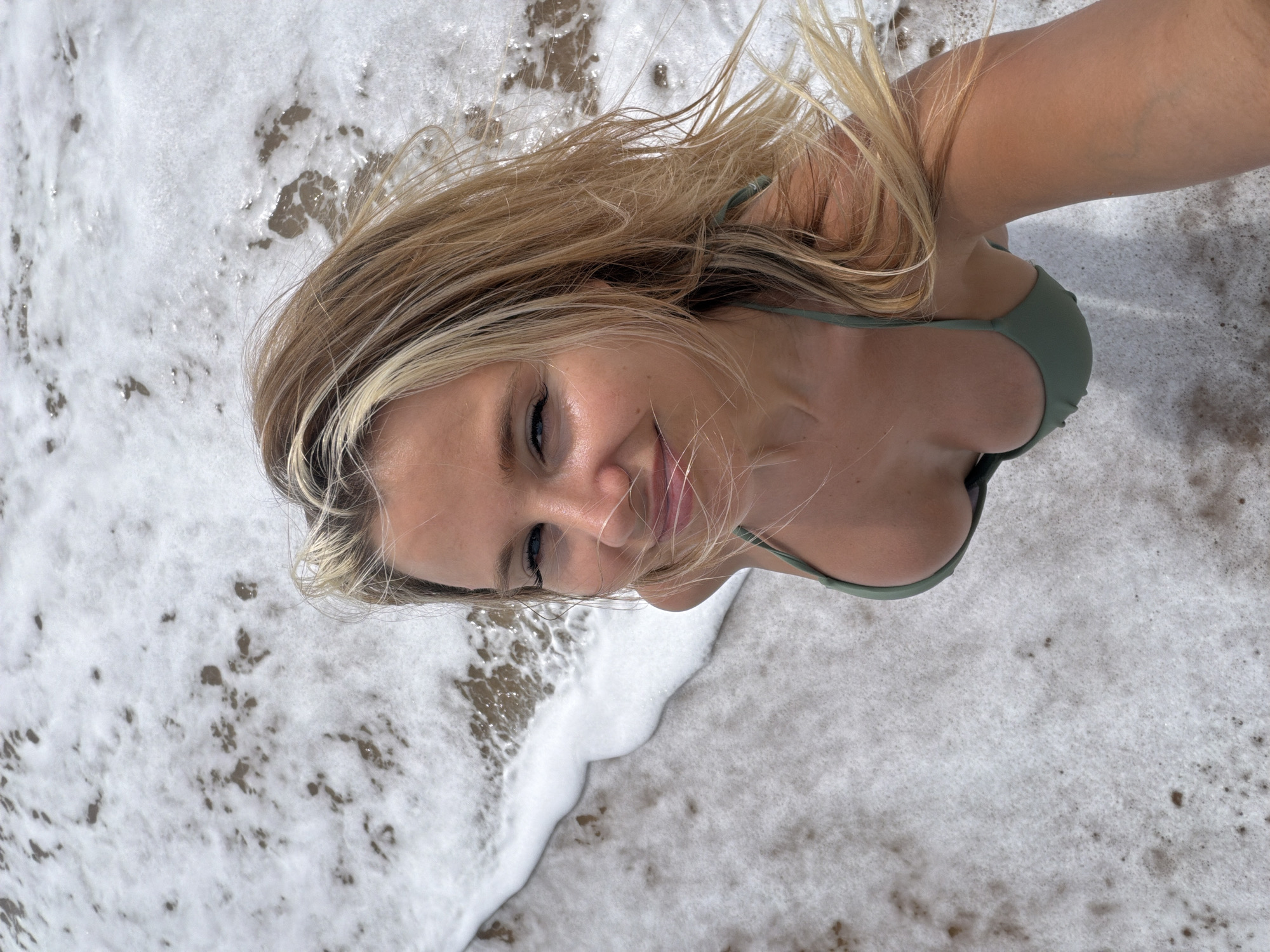 A young woman with blonde hair is posing for a selfie on a beach in Mai Khao, Thailand. She is wearing a green bikini top and is partially submerged in the ocean water, with waves lapping around her. The background is dominated by the ocean and the shore, with white foam and sand visible. The lighting suggests it is daytime.