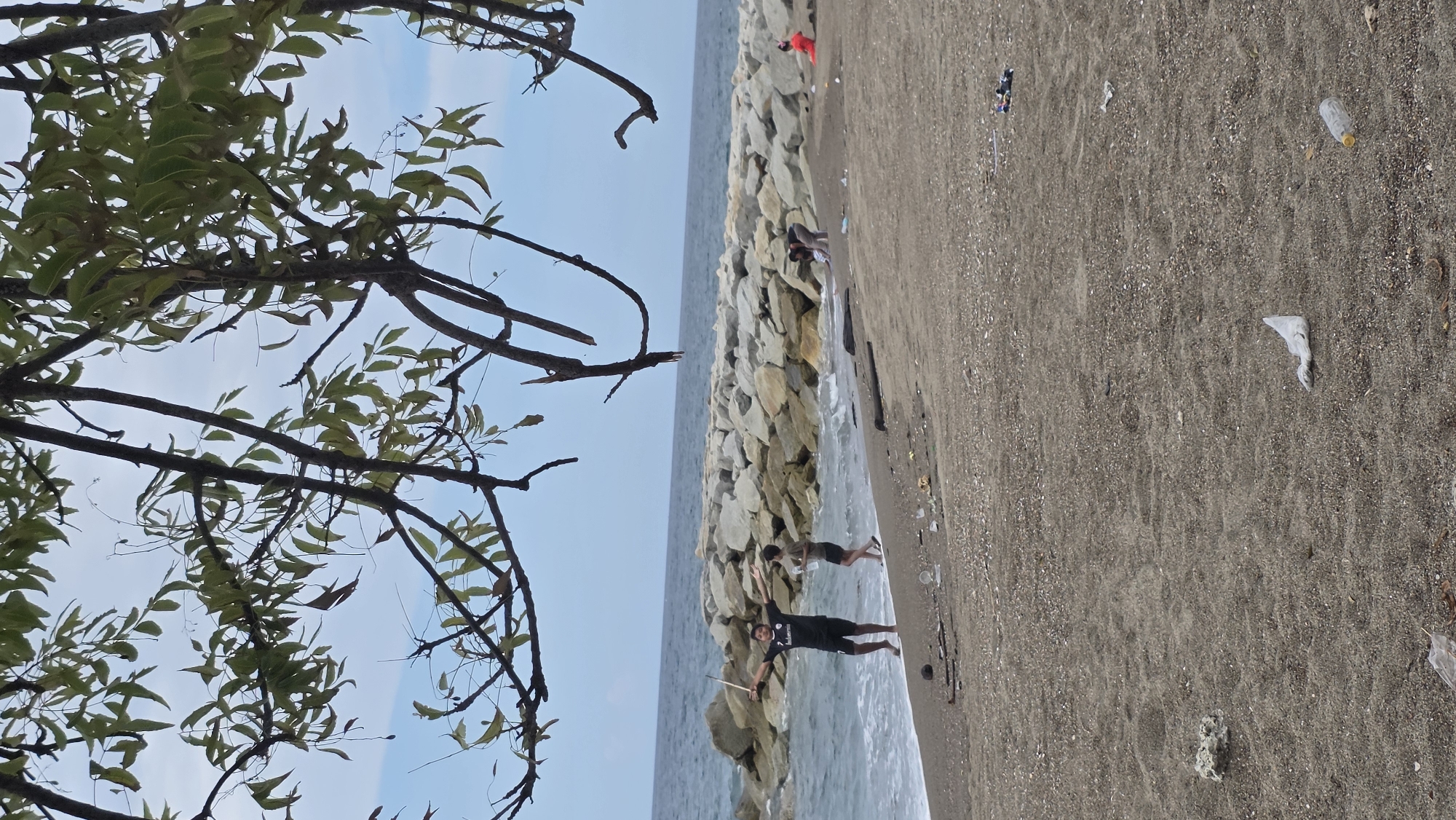 The scene depicts a beach environment in Lhokseumawe, Indonesia. The view includes the beach with grey sand, littered with plastic waste. A stone breakwater extends into the water, with waves washing against it. Several people stand on the beach near the water. A tree branch with leaves frames the left side of the image against a pale blue sky.