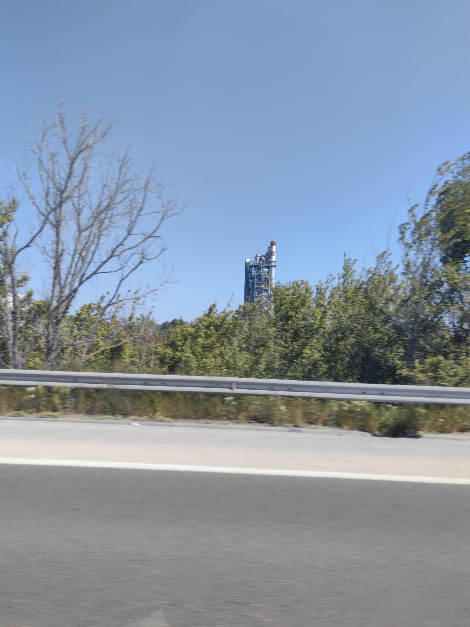 The image shows an industrial facility in Devnya, Bulgaria, viewed from a roadside. The facility features a tall, complex structure with red and white elements at the top, possibly a chimney or stack. This structure rises above a line of dense green trees and bushes, and a bare tree is visible on the left. A guardrail runs along the edge of the road, and the asphalt road surface is in the foreground. The sky above is clear and blue. The photo was taken from a moving vehicle.