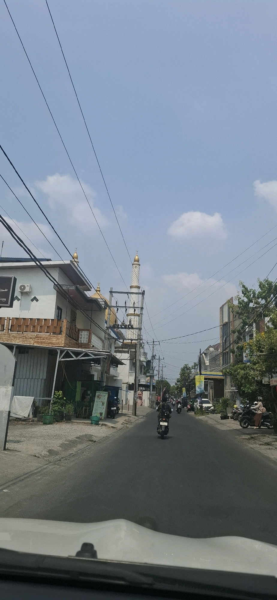 This is a photograph of a street in Pabelan, Indonesia. The view is from inside a vehicle looking forward. The street is paved and appears to be an urban or suburban setting.
On the left side of the street, there is a multi-story building with white walls and a brown wooden balcony. An air conditioning unit is visible on the second floor. Further down the street on the left, a mosque with a white minaret topped with a gold dome is prominent. The minaret has intricate white detailing. There are also power lines running across the sky.
On the right side of the street, there are several buildings, including one with a modern facade and signs. Trees with green foliage are visible, and some yellow flowers are present. There are also parked motorcycles and a few people.
Several motorcycles are in motion on the street, with riders visible. One motorcycle is in the foreground, heading away from the viewer. Another motorcycle is further up the street, also moving away. The sky is partly cloudy with a clear blue expanse. The overall impression is a typical day on a moderately busy street.