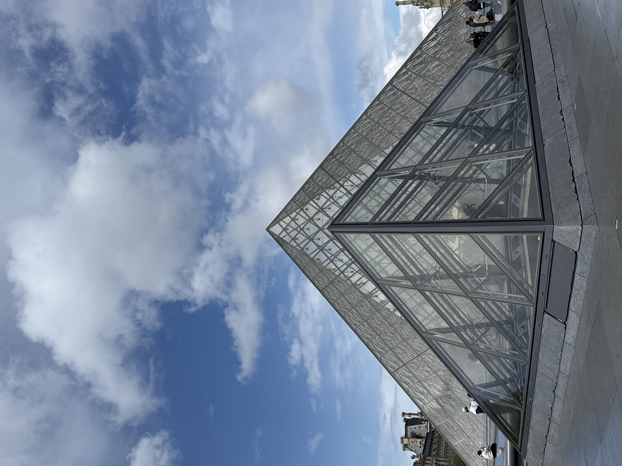 The image features the Louvre Pyramid in Paris, France, from a low-angle perspective. The pyramid's glass and steel structure dominates the right side of the frame, contrasting with the blue sky filled with scattered clouds. A few people are visible on the perimeter of the pyramid and reflect on the glass, suggesting it is a place for gathering. The surrounding pavement and parts of the historic Louvre Palace are visible.