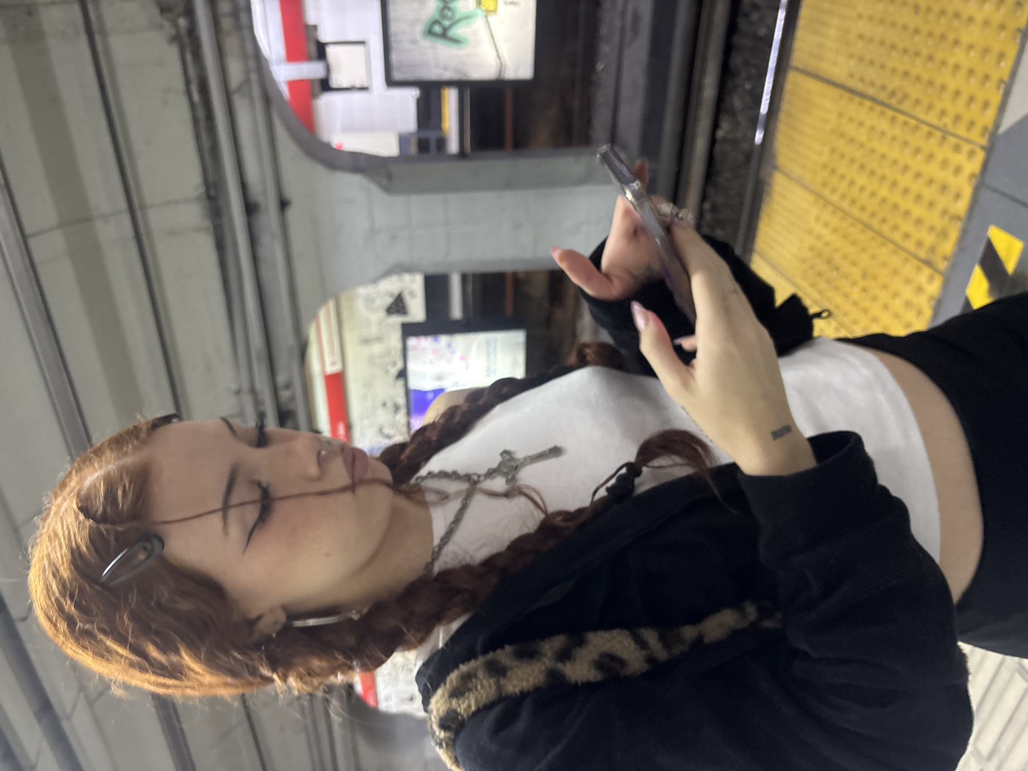 A young woman with reddish-brown hair in braids stands on a subway platform. She is looking at her cell phone. She is wearing a white tank top, black jacket, and a necklace with a cross pendant. The platform has yellow tactile paving along the edge. Signage and architectural details suggest that the location is a subway station in Buenos Aires, Argentina.