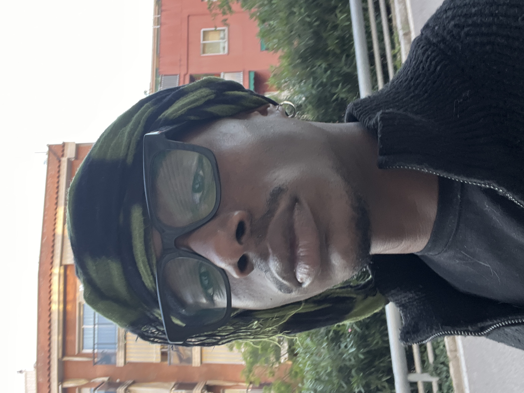 A close-up, eye-level portrait of a Black man wearing glasses and a patterned head covering. He is positioned slightly off-center. The background features a building with windows, suggesting an urban environment. In the far background, foliage of trees. The scene appears to be set outdoors, possibly on a street or balcony. Given the architectural style of the building, this image might be taken in the city of Barcelona, Spain.