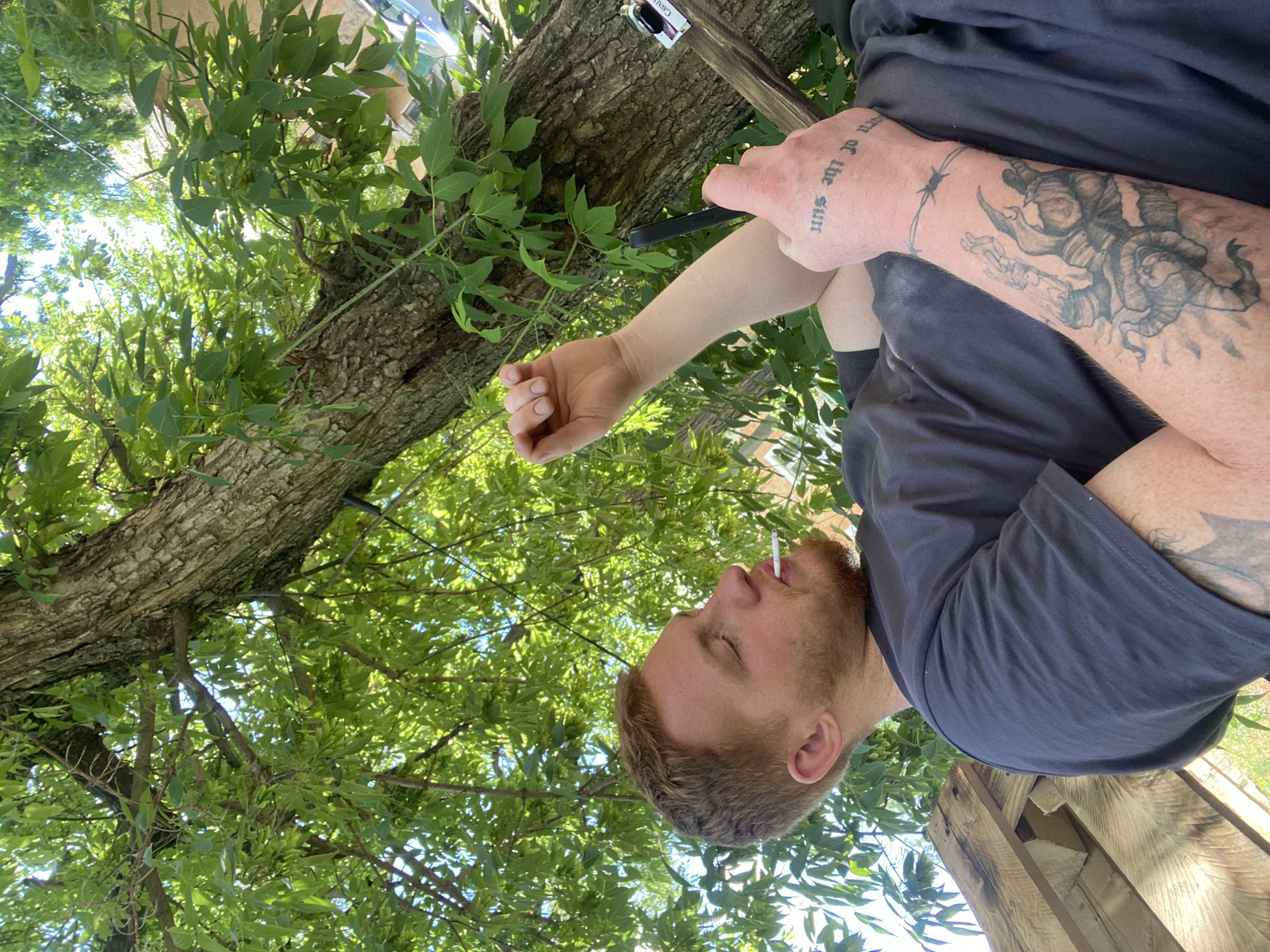 An outdoor, low-angle shot features a man with red hair and a beard sitting near a tree. He is wearing a dark gray t-shirt and has a cigarette in his mouth. His left arm is visible, displaying several tattoos, including barbed wire around his wrist and a large, intricate design on his forearm. He is holding a dark smartphone in his right hand, which is raised. The background consists of lush green leaves and tree branches, with sunlight filtering through. The man appears to be in a relaxed or contemplative pose. There are no other people or animals visible in the frame. The scene suggests a casual, possibly private, outdoor setting.