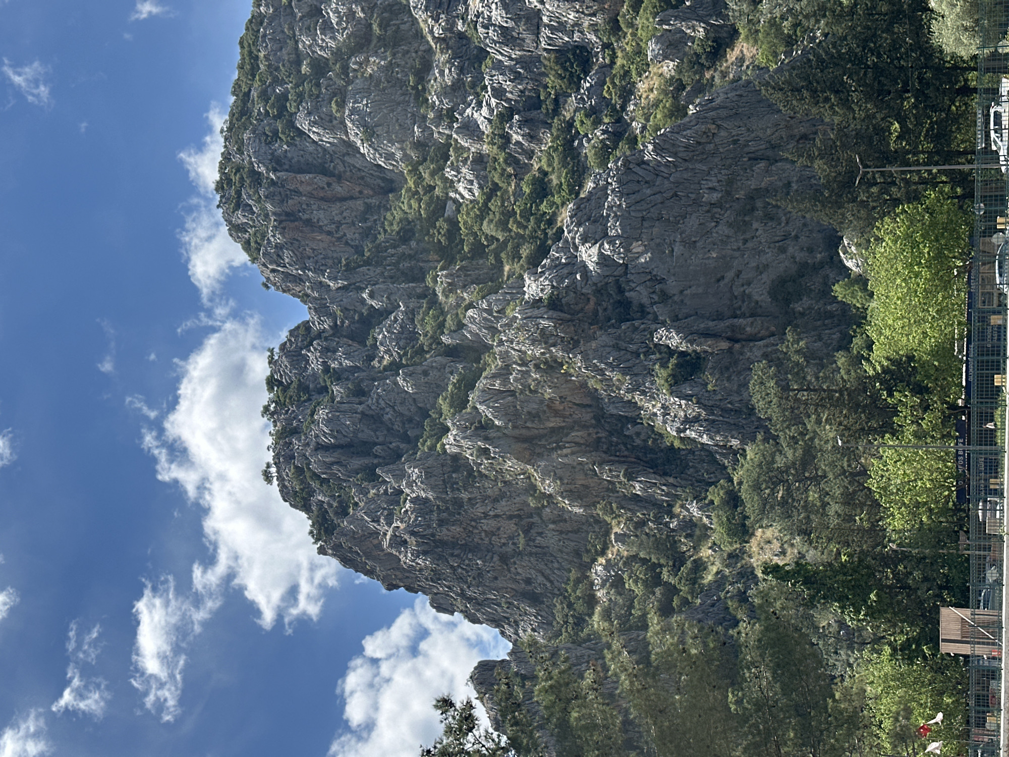 The image captures a rocky mountain landscape, possibly near Kumluca, Turkey, given the prompt's specification. The mountain is predominantly gray with sparse green vegetation clinging to its slopes and crevices. To the left, a blue sky is visible with scattered white clouds. A segment of trees and perhaps a fence or structure is seen to the right, partially obscuring the mountain's base. The composition appears to be taken from a low angle, emphasizing the height and ruggedness of the mountain.
