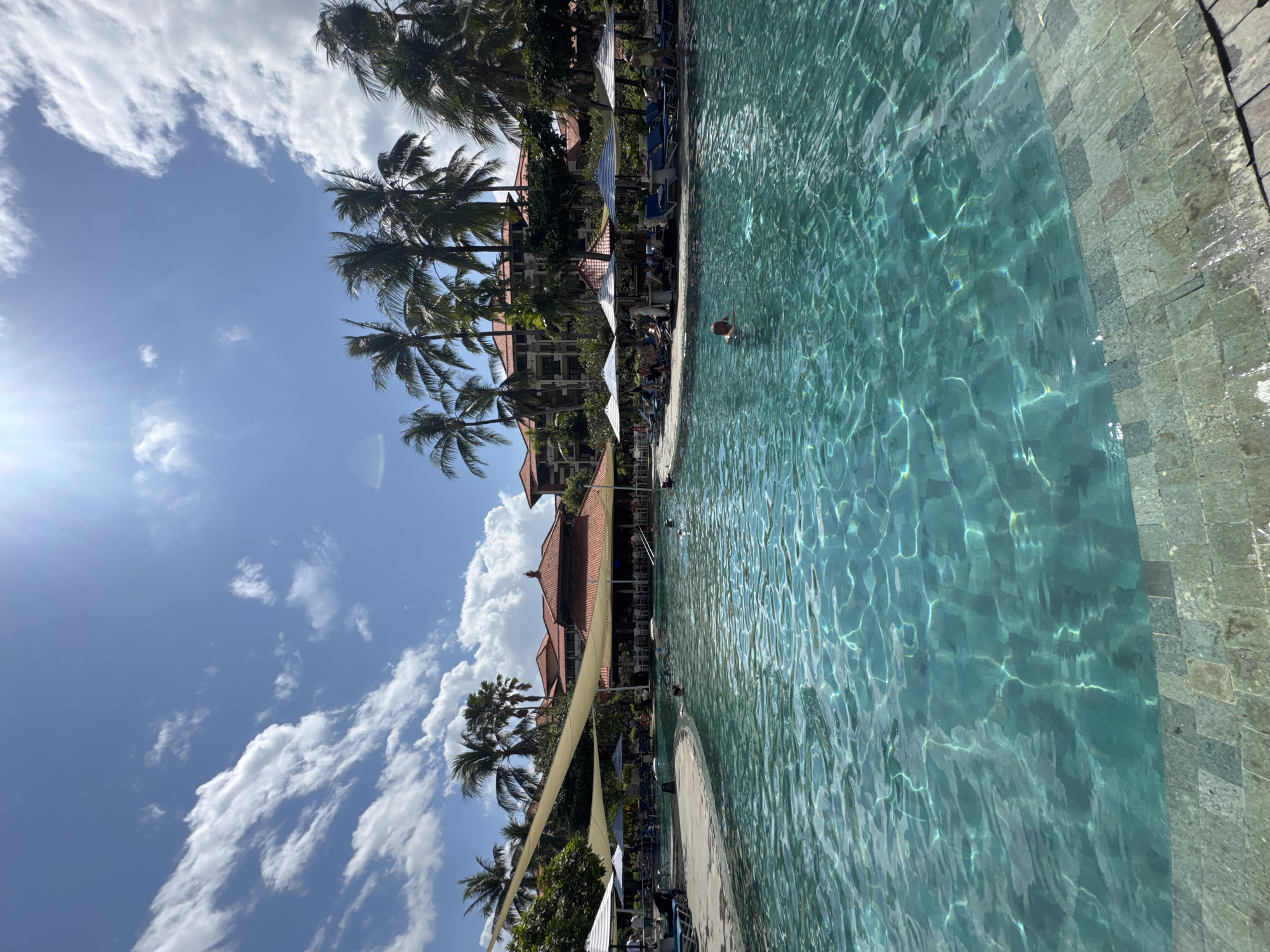 The image depicts a large, clear turquoise swimming pool with sunlight glinting off the surface, creating a shimmering effect. The pool is surrounded by a stone-tiled edge. In the background, there is a building with a terracotta-tiled roof, a bar area, and numerous palm trees. Several people are visible, some swimming in the pool, while others are seated on lounge chairs under umbrellas or the shade of canopies on the poolside. The sky is blue with scattered white clouds, and the overall scene suggests a tropical resort environment. This location is likely within the city of Kuta Selatan, Indonesia, a region known for its resorts and beaches.