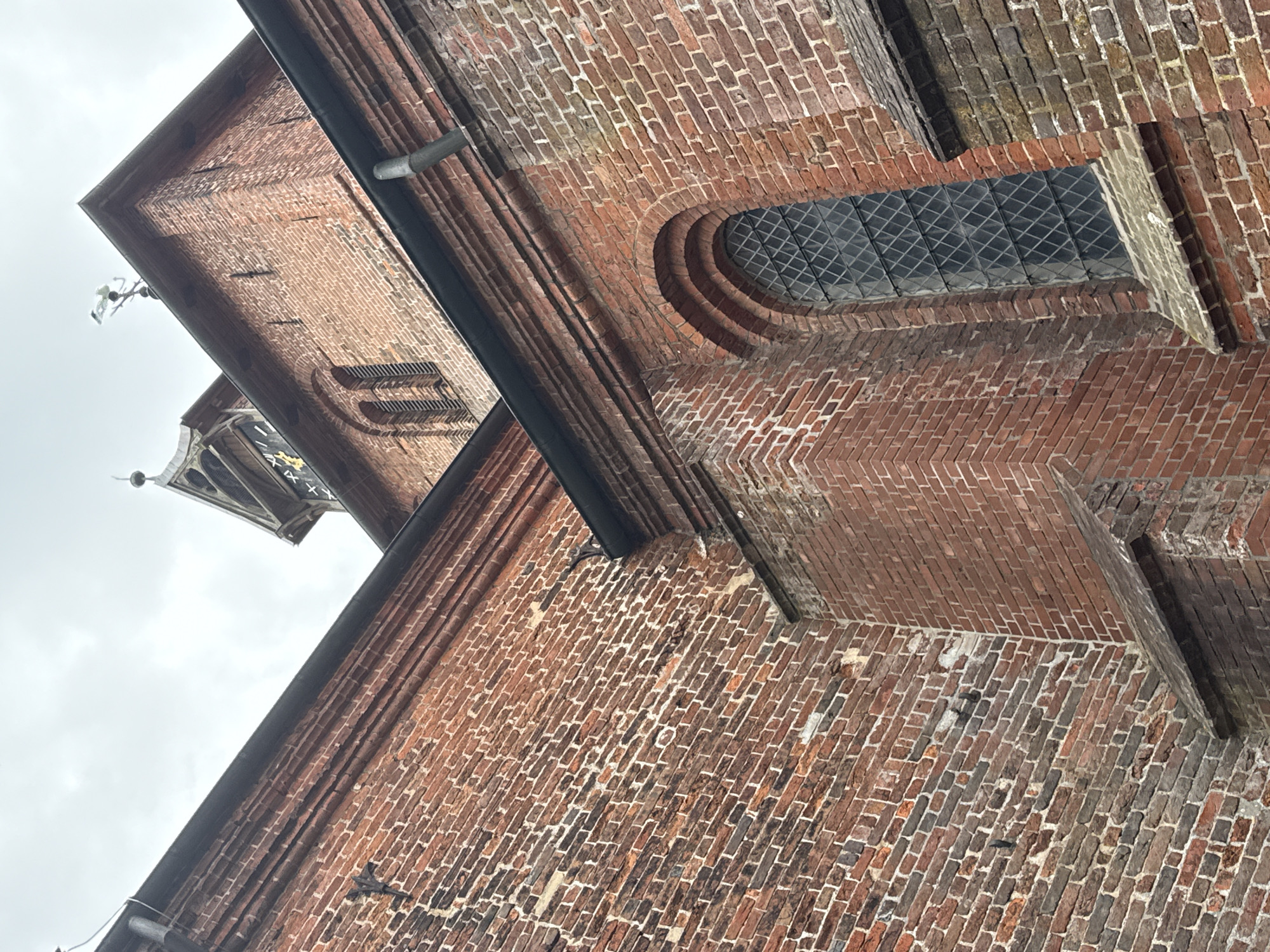 This is a low-angle, full-shot photograph of the exterior of a brick building, likely a church or historical structure, in Bassum, Germany. The image is vertically oriented and appears to have been taken on an overcast day, contributing to the muted natural light.
The primary subject is the intricate brickwork, which varies in color from deep red to shades of brown and gray, indicating age and possibly historical repairs. The building features architectural elements such as a pointed window with a diamond-patterned glass, an arched window recess, and a downspout. The sharp contrast between the dark downspout and the lighter brickwork draws attention. In the upper left portion of the image, a section of a tower with decorative elements and what appears to be a clock face with Roman numerals is visible against the bright, cloudy sky. Faint signs of decorative ironwork or metal fixtures can be seen attached to the brick facade.
There are no people or discernible activity taking place in the image; it is a static architectural study. The overall mood is somber and historical, emphasizing the age and solidity of the structure. The setting is clearly urban or semi-urban, given the architectural style and the presence of what seems to be a prominent tower. The cloudy weather suggests it could be any time of day, but the lighting is not harsh, implying it is not direct midday sun. No legible text is visible in the image, apart from the Roman numerals on the clock face which are partially obscured.