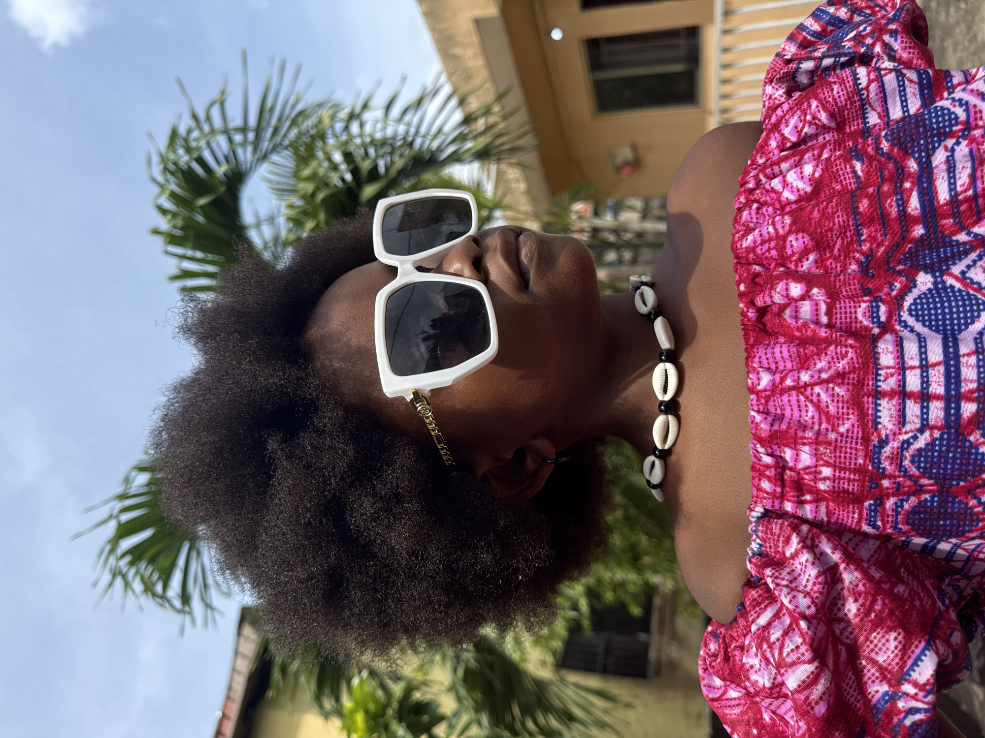 A medium shot depicts a dark-skinned person with afro-textured hair, wearing white sunglasses and a necklace with shells, a gold earring chain, and a pink and blue patterned top. They are looking upwards against a backdrop of a palm tree and a multi-story building with windows, under a partly cloudy sky. The location appears to be Ado, Nigeria.