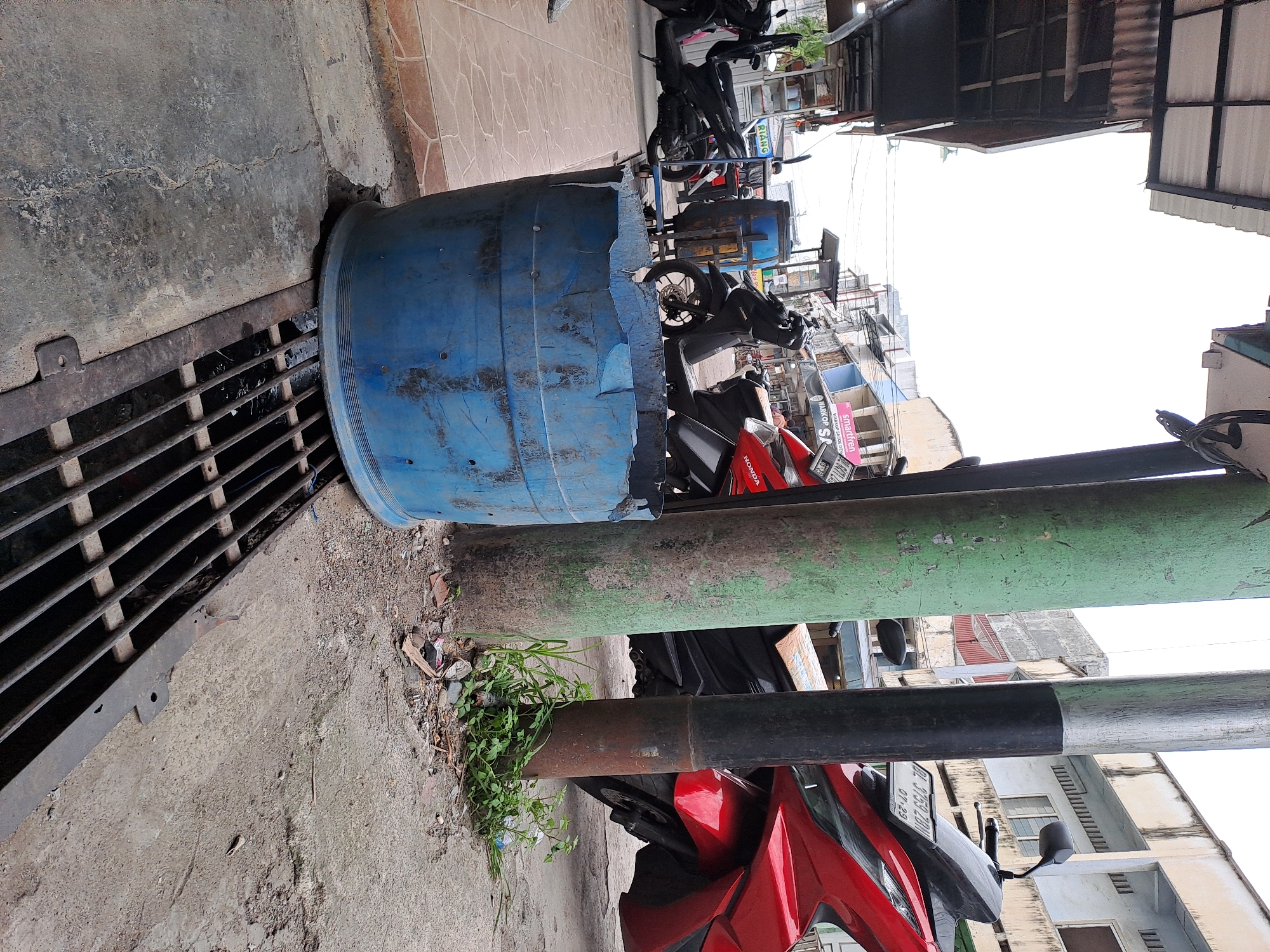 The image depicts a street scene in Bandarbireuen, Indonesia. In the foreground, a large, dented blue plastic barrel sits partially over a metal grate, which appears to be a drainage cover. To the right of the barrel, a red motorcycle is parked, partially obscured by a black pole. Behind the motorcycle, another black motorcycle is visible. The background shows a row of buildings with some shops, indicated by signage. The overall environment is urban, with paved surfaces and a sense of everyday street life. The sky is overcast.