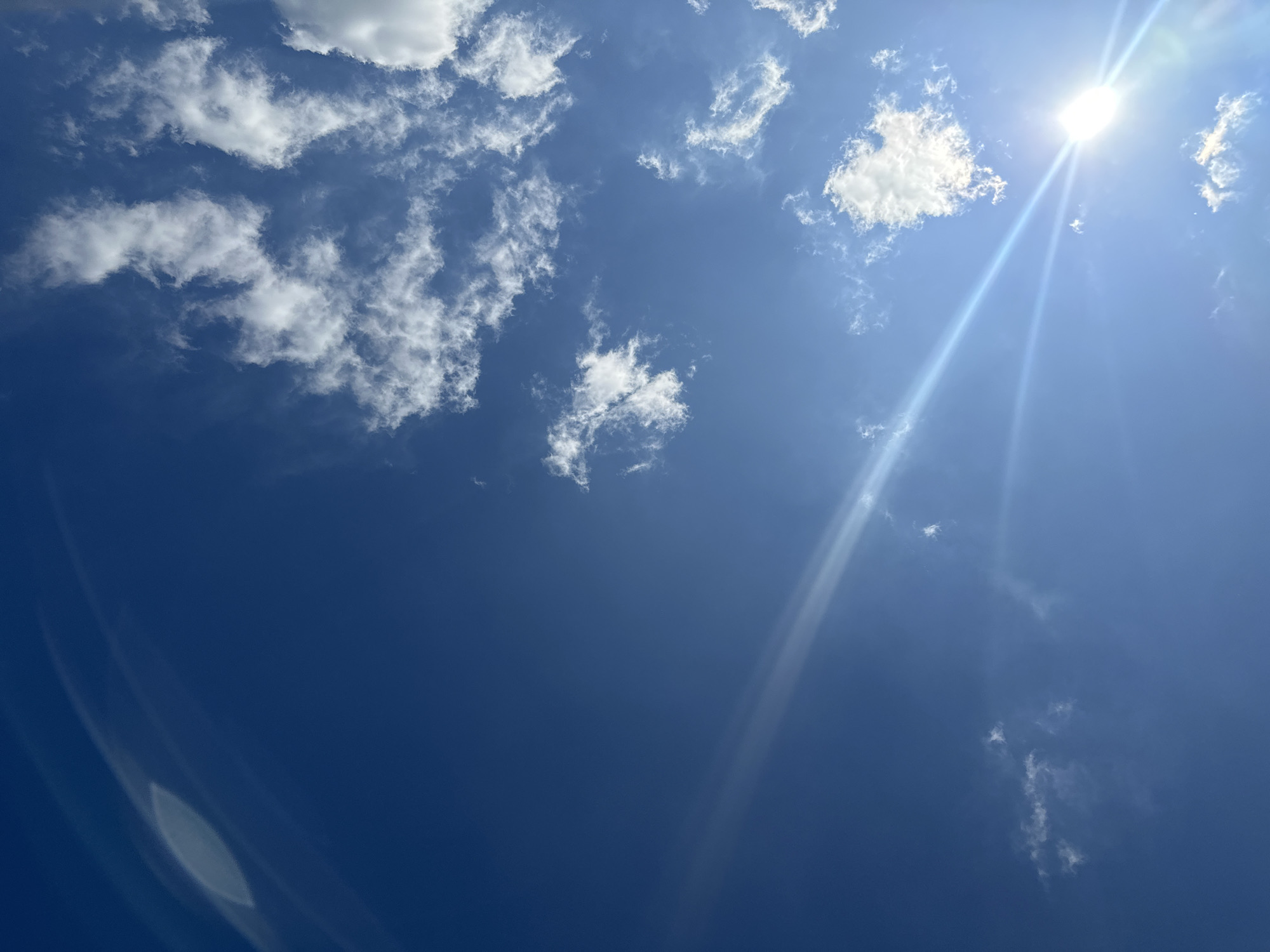The image shows a bright blue sky with scattered white clouds. A prominent sunbeam streaks across the sky from the lower right corner, illuminating the clouds and creating lens flare effects. The scene is characteristic of an outdoor environment, likely captured during daylight hours. The provided location, Plesetsk, Russia, is a city known for its spaceport, but this image provides no specific clues to that context. The image contains no people, subjects, objects, or actions. The environment is a sky with clouds.