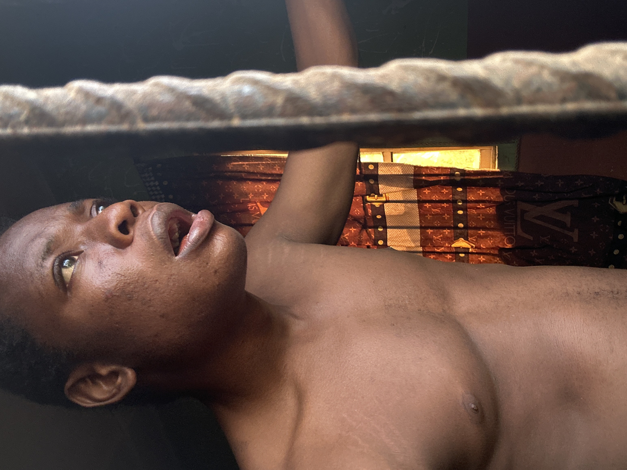A shirtless man lies on his back, looking upward. He is positioned beneath a dark horizontal object, possibly a beam or ledge. A window with light shining through is behind him. A Louis Vuitton patterned textile hangs in the background near the window. The location is Unknown, Unknown.