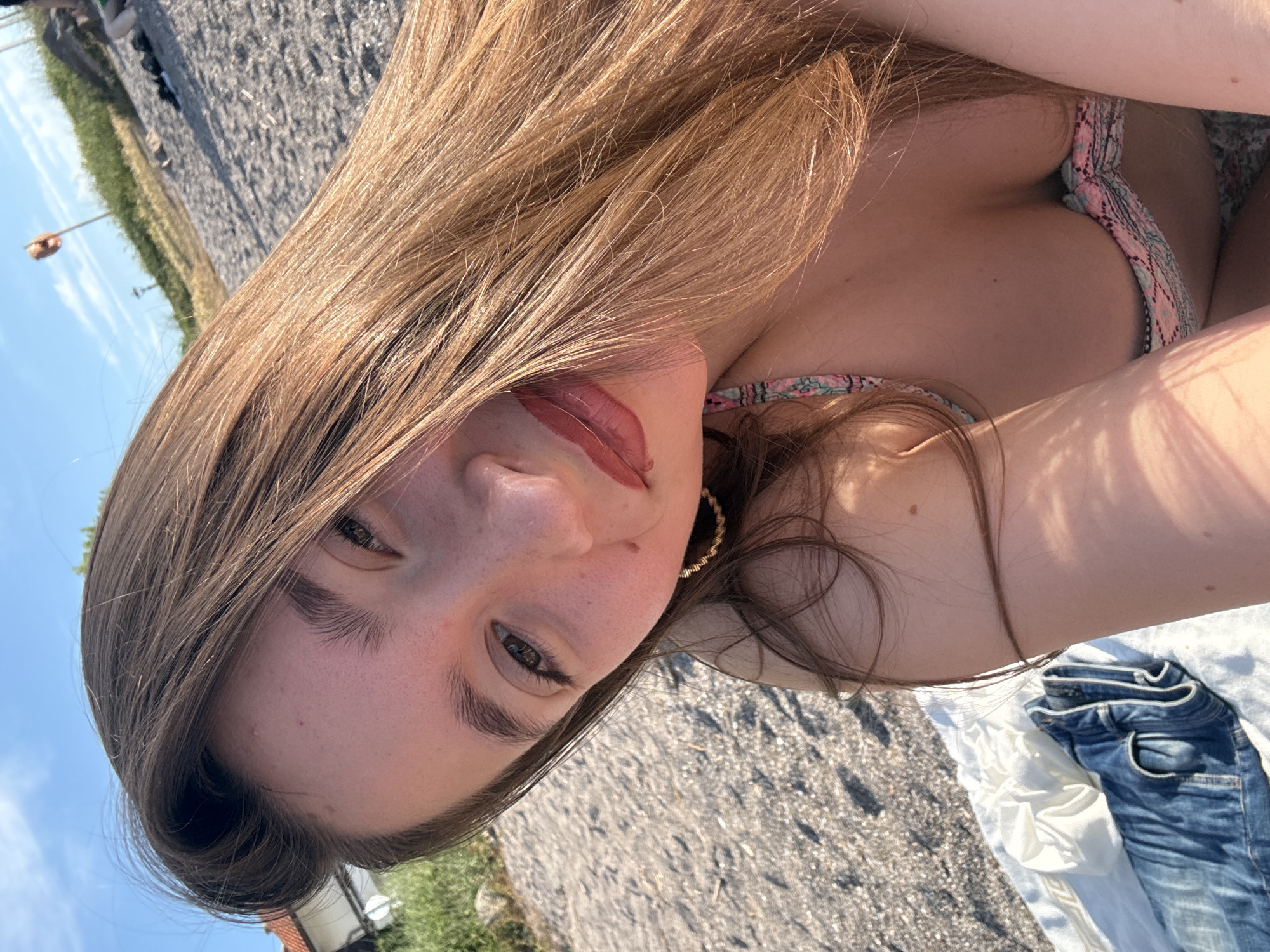 The media file is a portrait of a young woman lying down outdoors on a sunny day. She has long, light brown hair that partially covers her face. Her eyes are open and looking towards the camera, and she is wearing subtle makeup. She has on a bikini top with a patterned design. In the background, there is a grassy area, a sandy or gravelly ground, and a clear blue sky. A pair of blue jeans is folded and placed on what appears to be a white sheet or towel to her right. The lighting suggests it is daytime, likely late afternoon due to the angle of the sun casting shadows. The overall mood is relaxed and casual, possibly at a beach or park. There is no visible text in the image.