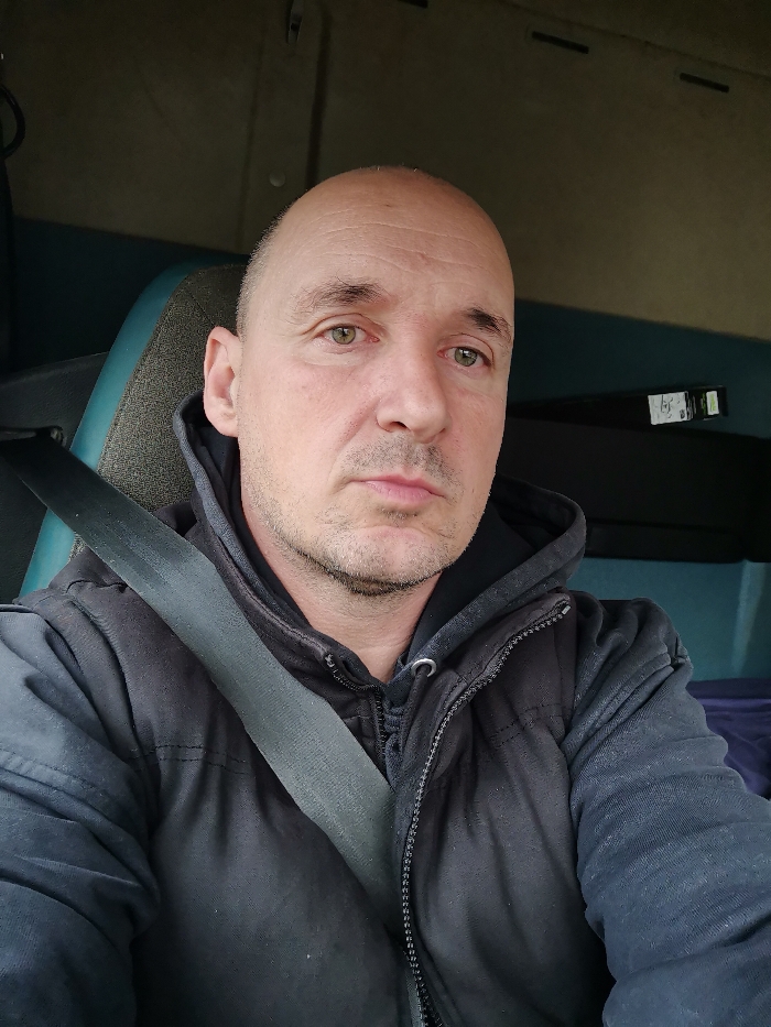 The image displays a male subject, appearing middle-aged, seated inside the cabin of a vehicle, specified as located in Pchelarovo, Bulgaria. The individual has a bald head, light stubble on his chin and upper lip, and greenish-hazel eyes, maintaining a neutral expression while looking directly at the camera. He is wearing a dark, possibly black, hooded garment beneath a dark, quilted, zipped-up vest or sleeveless jacket. A dark gray seatbelt is positioned diagonally across his chest and left shoulder. The vehicle's interior shows a seat with a dark teal frame and a textured grey fabric insert. Above, the roof lining is a light neutral color. To the subject's right, a dark background panel is visible, upon which a small rectangular device with faint white text/graphics and a green indicator light is mounted. Below this, a dark blue or purple fabric item is present.
